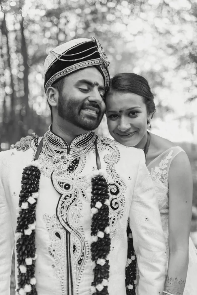 A couple dressed in traditional Indian wedding attire, smiling and embracing outdoors with a blurred background of trees.