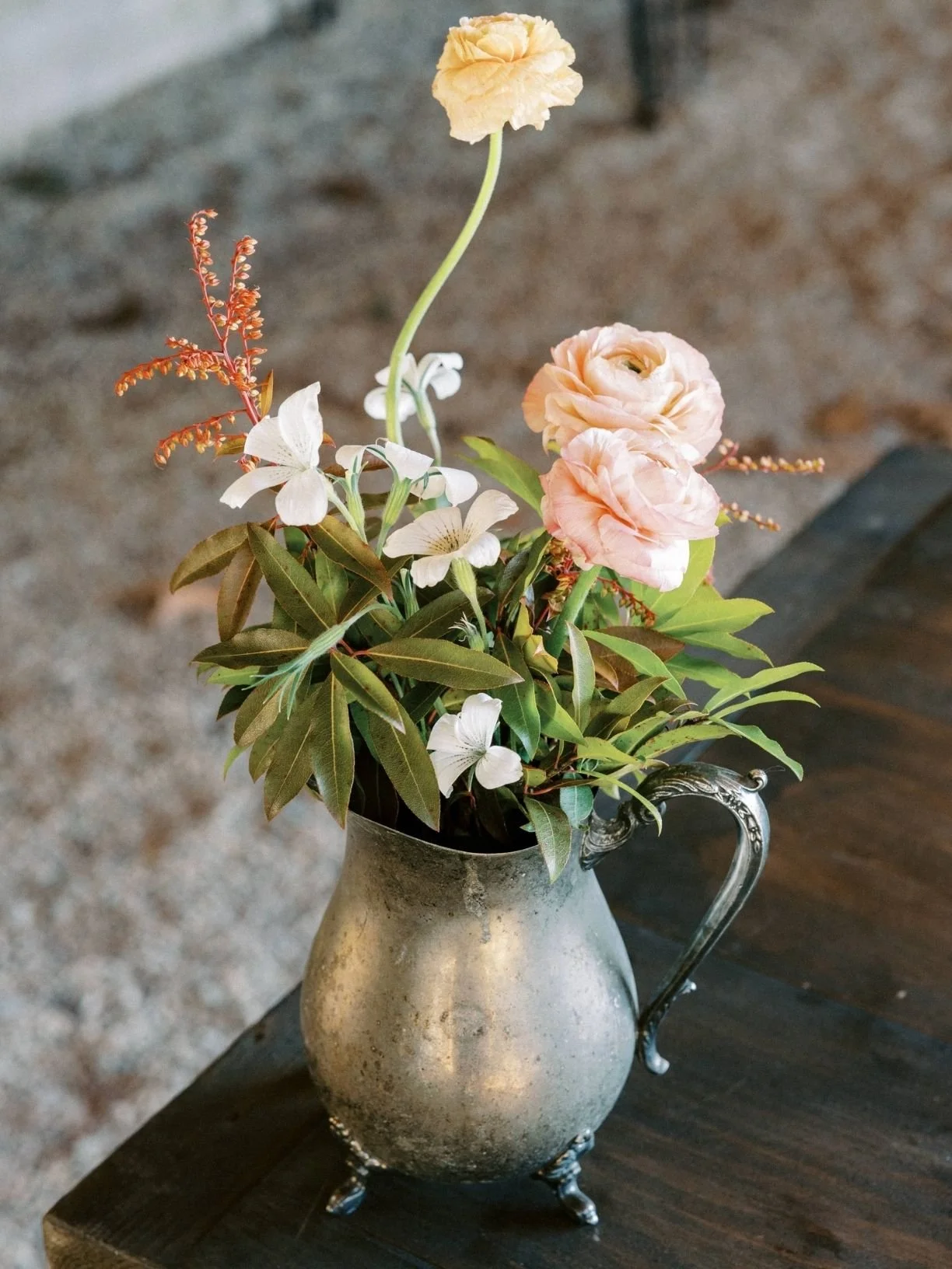 A silver metal pitcher with a floral arrangement of pink and white flowers and green leaves, placed on a dark wooden surface.