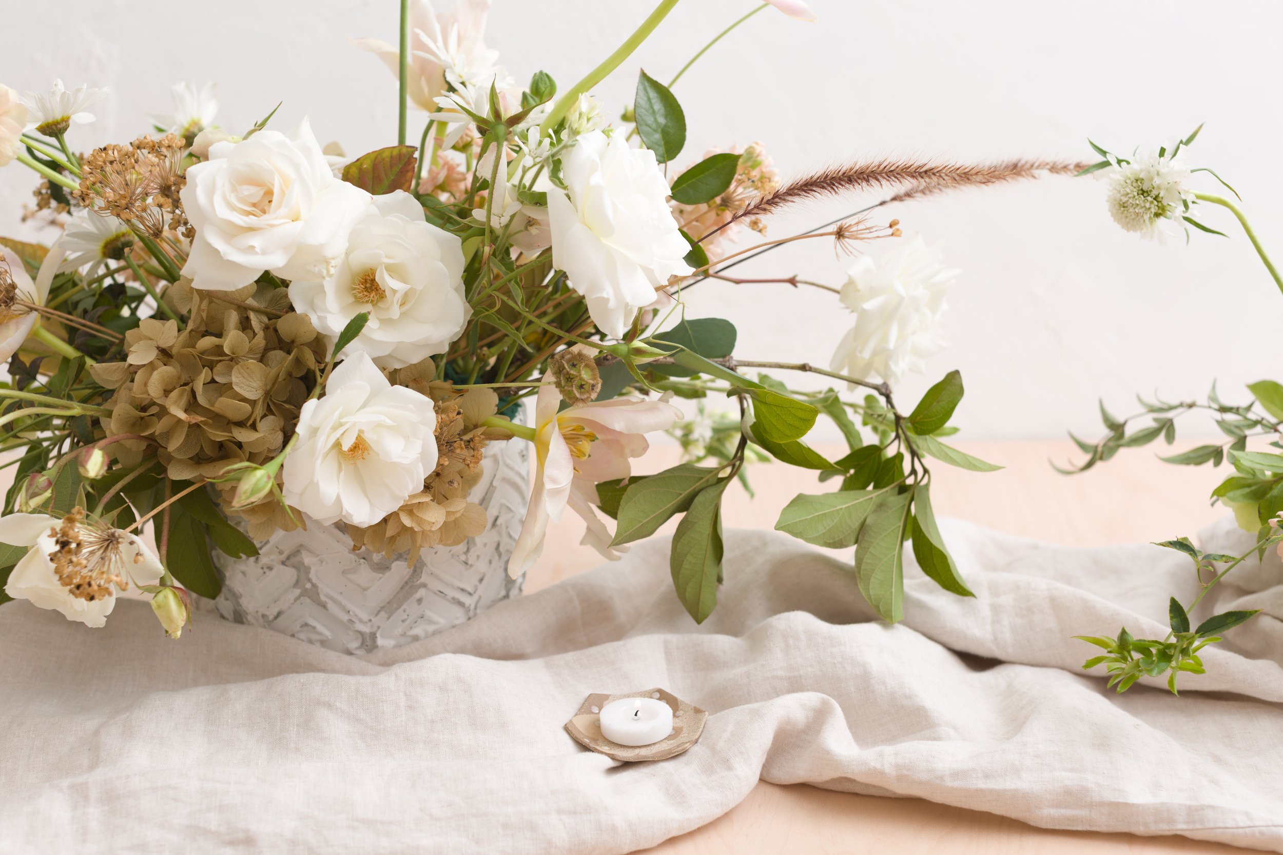 A floral arrangement of white and beige flowers with green leaves in a textured white vase on a linen cloth, with a small tealight candle holder nearby.