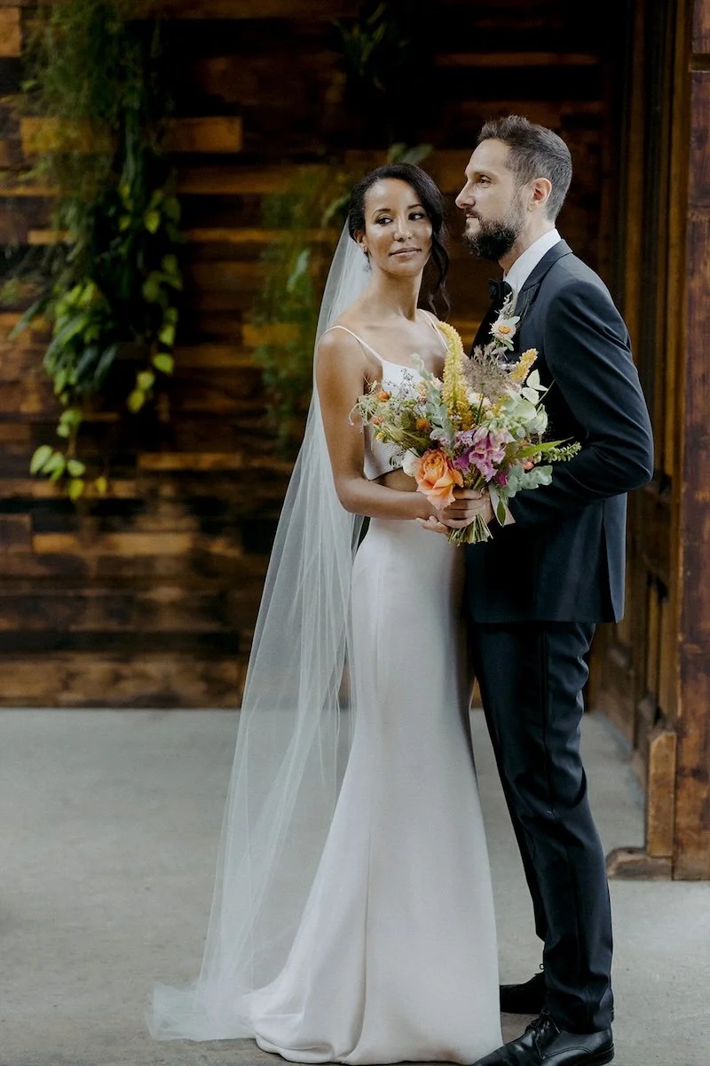 Bride and groom standing indoors, the bride holding a bouquet, wooden wall with greenery in the background.