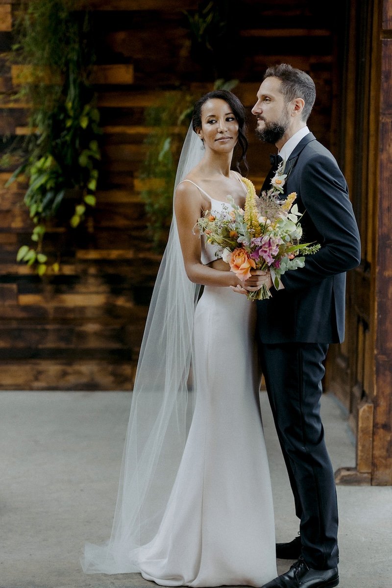 Bride and groom standing indoors, the bride holding a bouquet, wooden wall with greenery in the background.