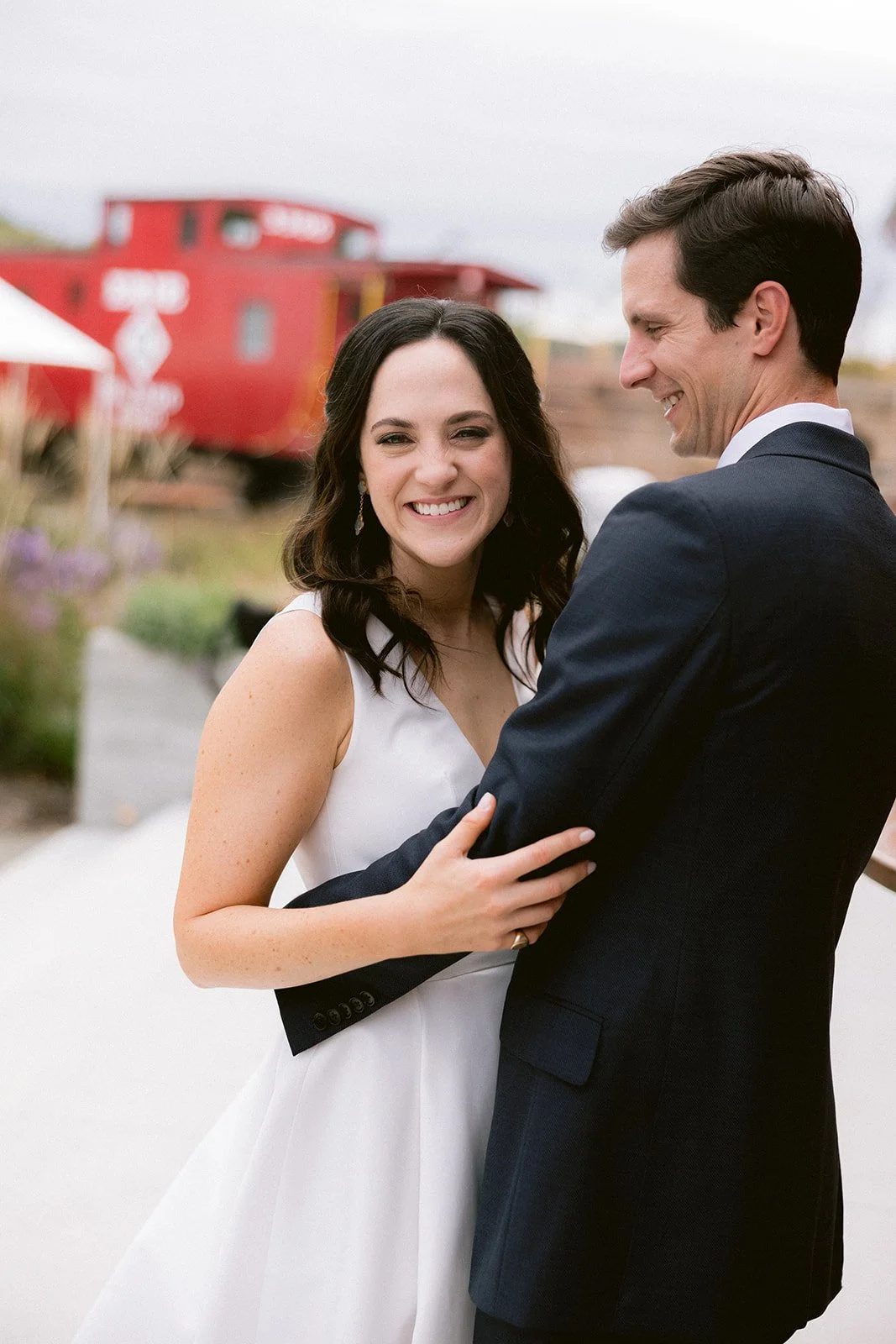 A couple in wedding attire smiling, with a red train in the background.
