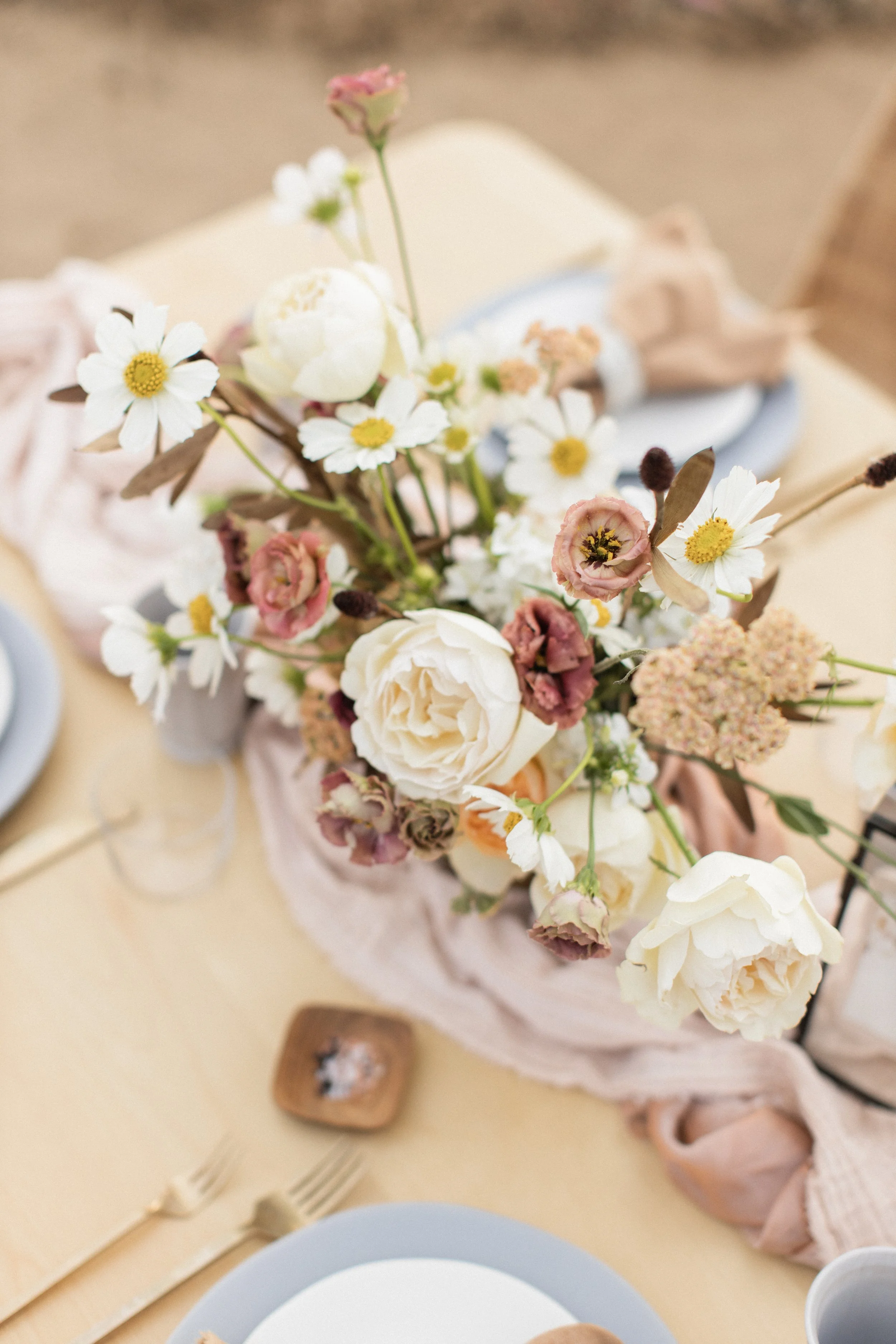A floral centerpiece with white and blush pink flowers on a dining table, decorated with a blush pink fabric runner, set for a meal with plates, cutlery, and a small wooden dish underneath.