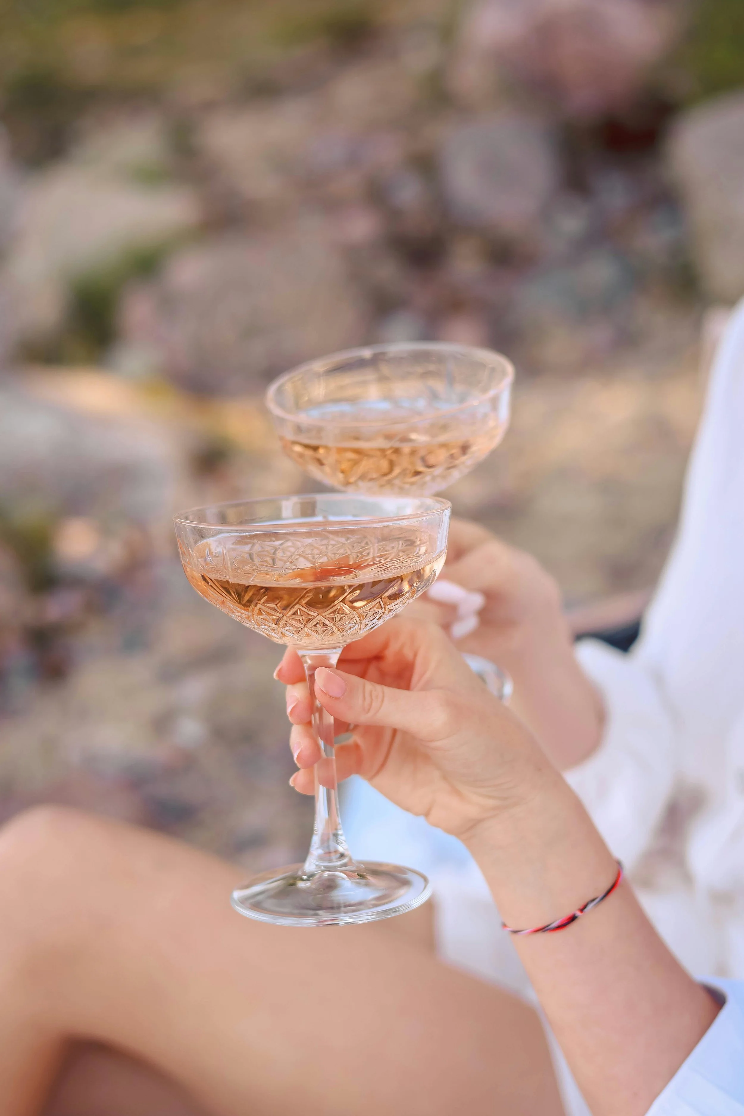Person holding two coupe glasses with pink bubbly beverage, with a rocky outdoor background.