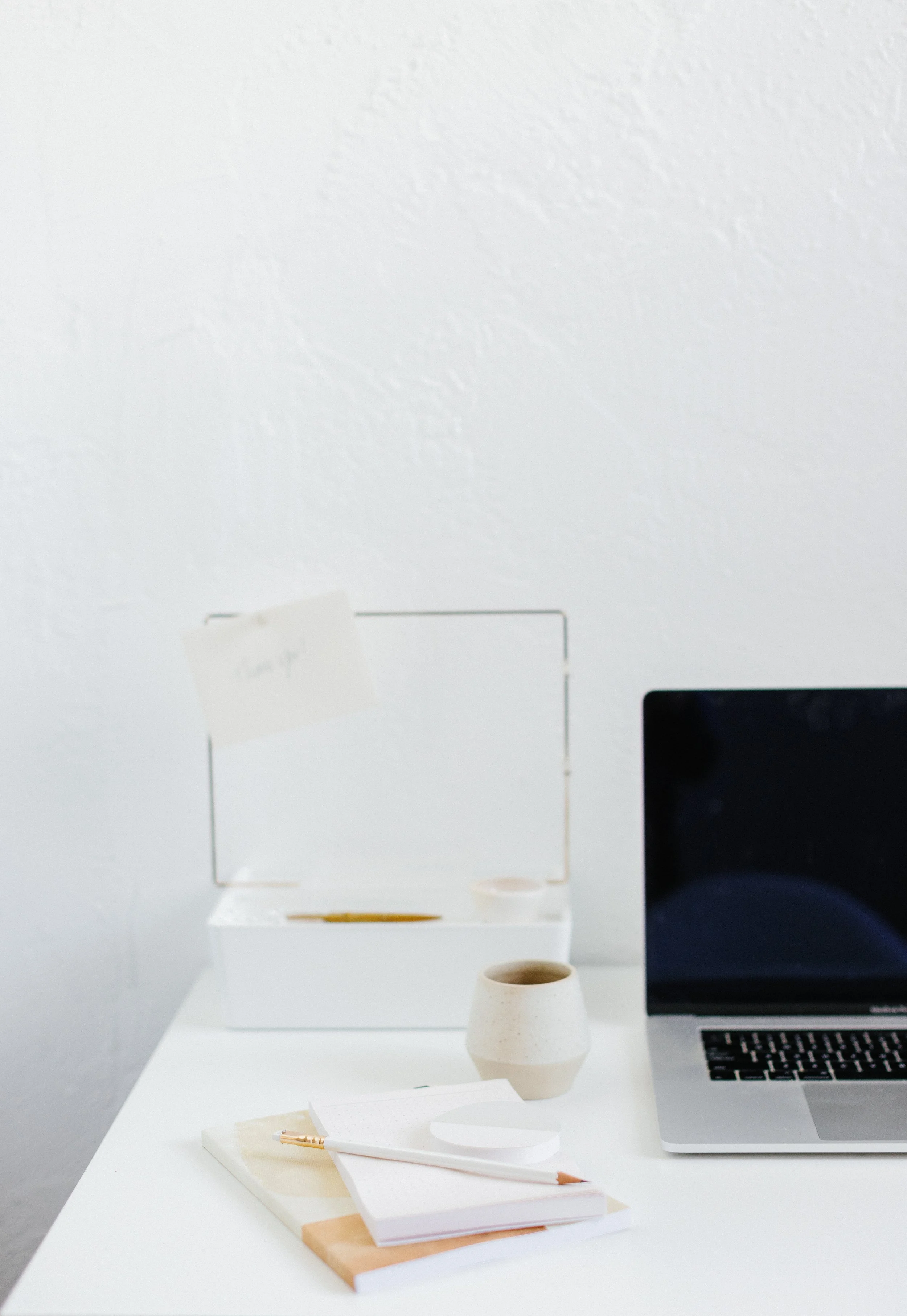 A minimalist workspace with a closed laptop, a white notepad with a pencil, a small beige ceramic cup, and an open white box with a mirror, all set on a white desk against a white wall.