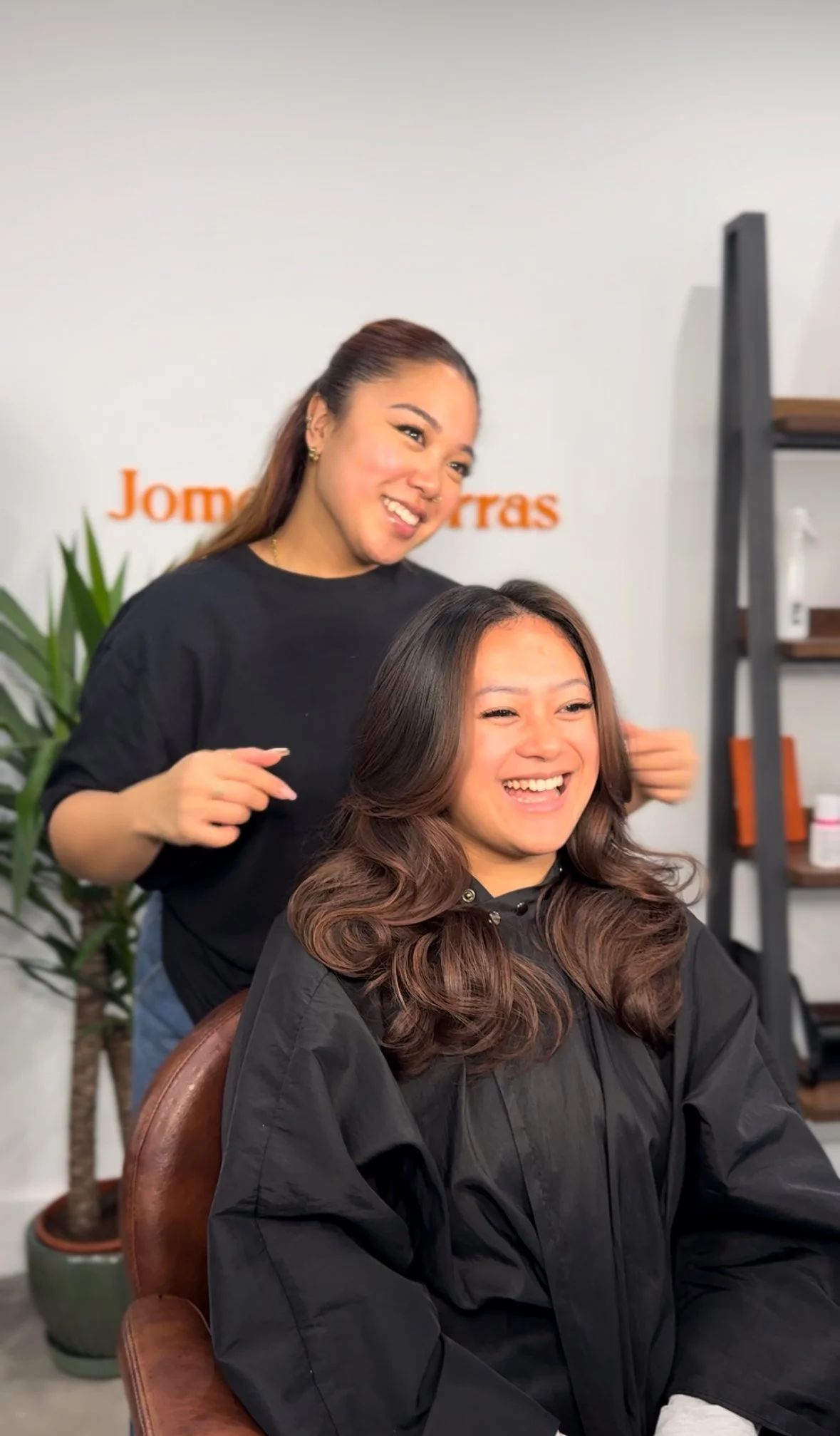 A woman with long, wavy brown hair smiling in a black salon cape, getting a haircut from a hairstylist in a salon.