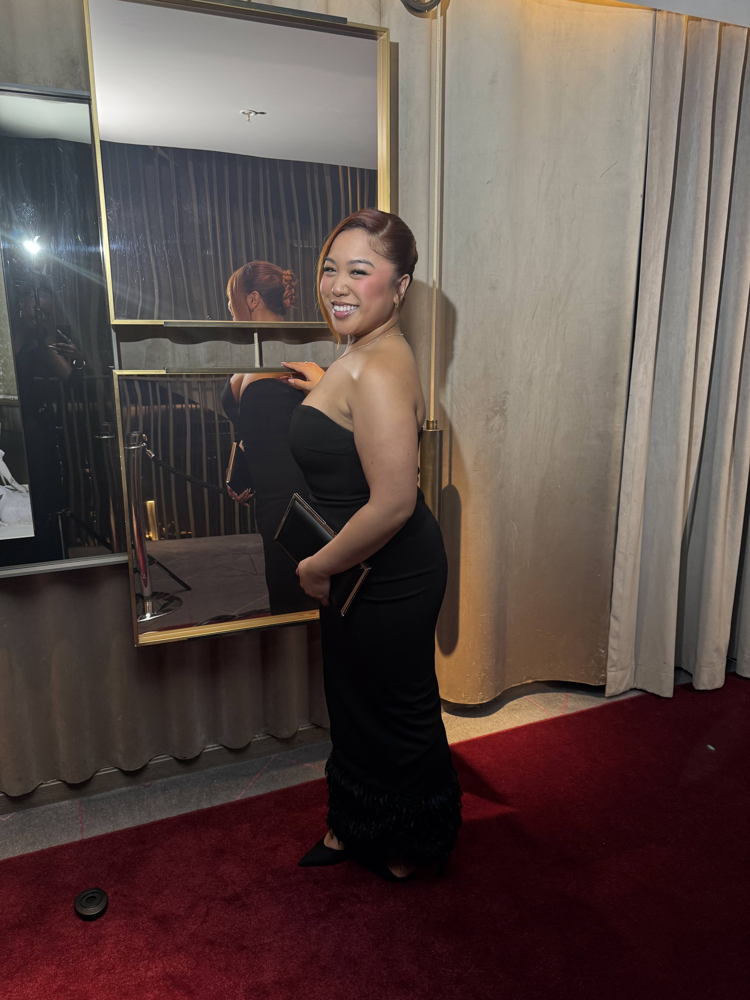 A smiling woman wearing a strapless black evening gown with feathered hem, holding a black clutch, standing in front of a mirror in a hotel or event setting.