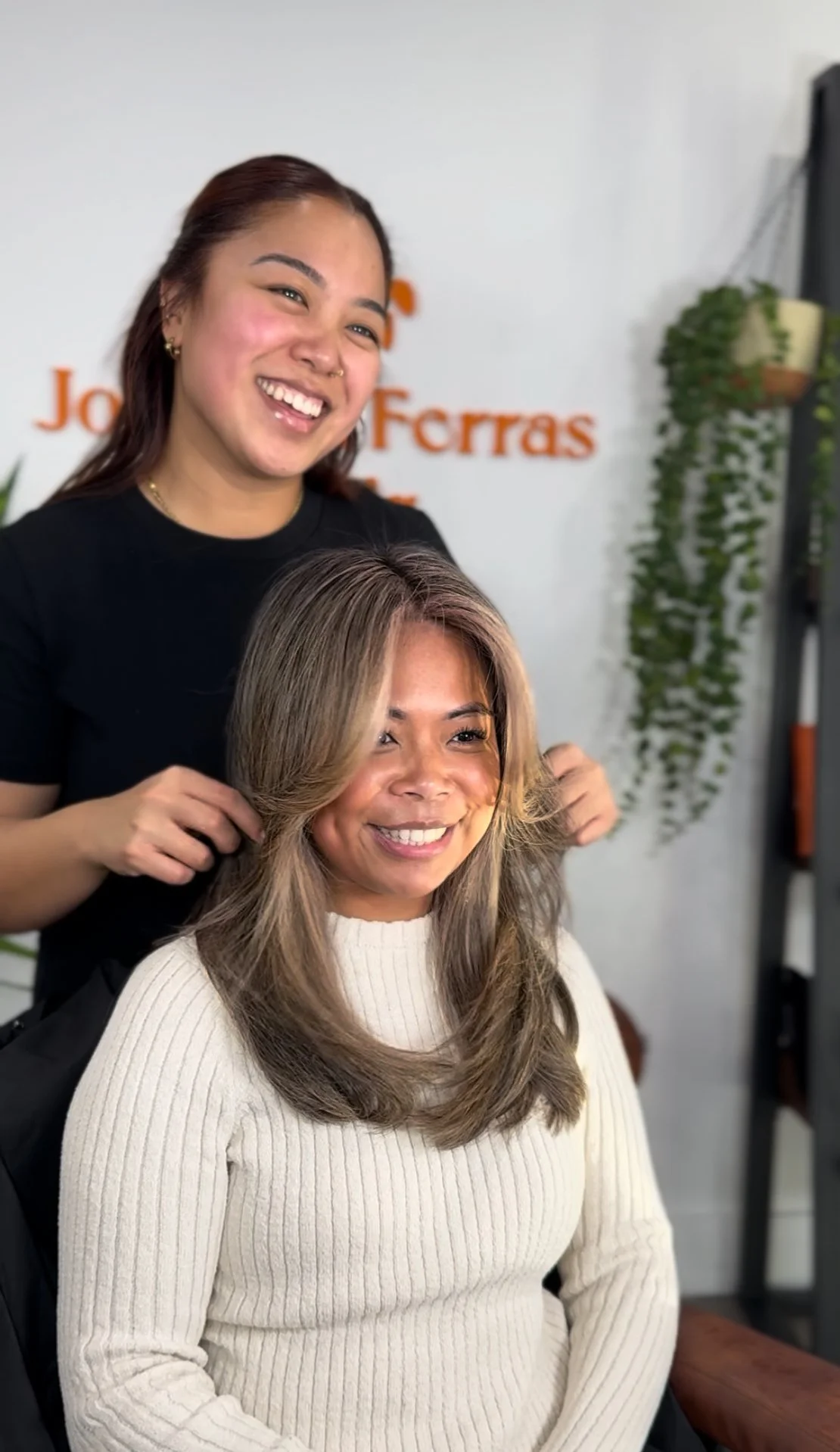 A hairstylist is working on a woman's hair in a salon, both are smiling.
