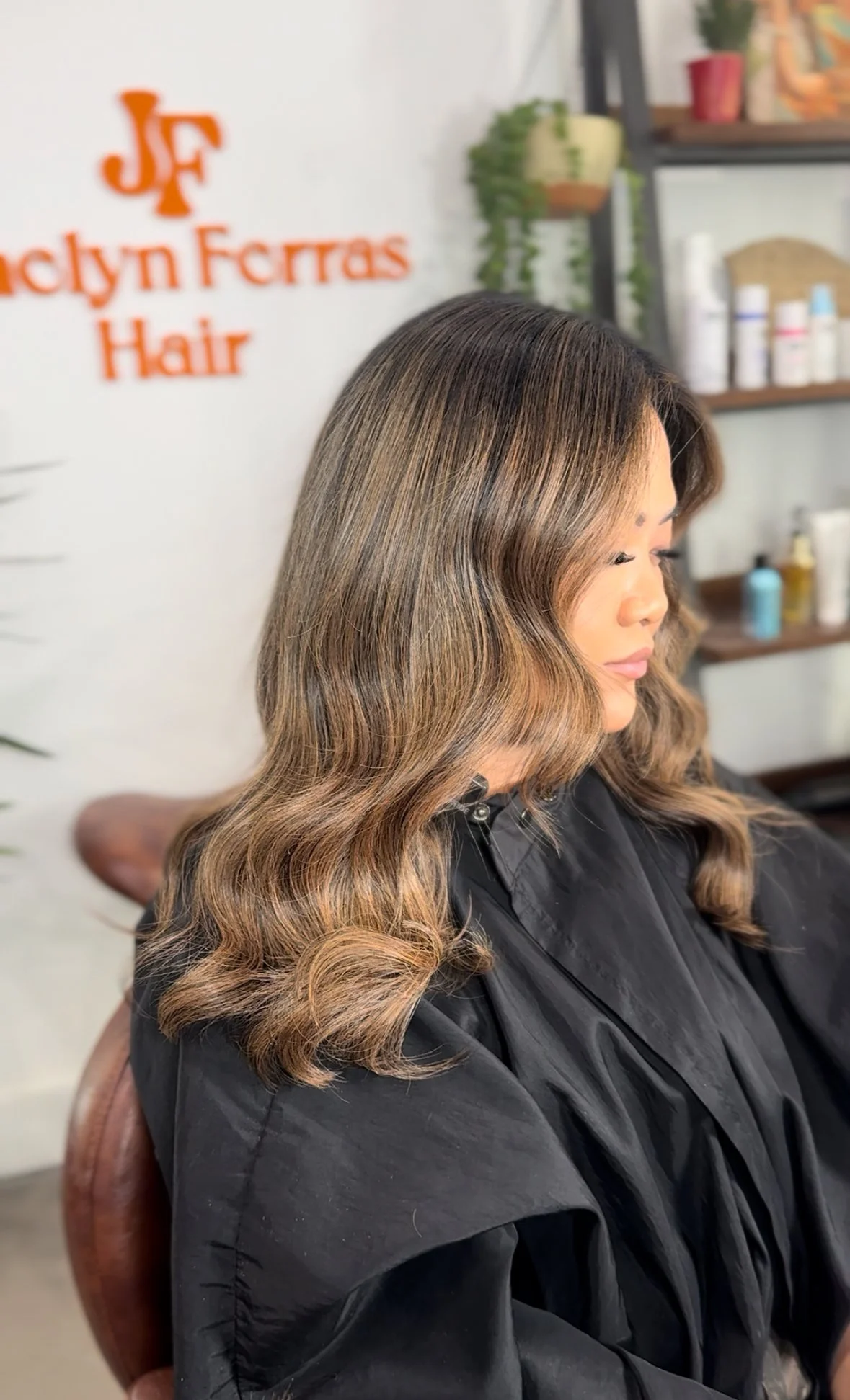 A woman with long, wavy hair with blonde highlights sits in a hair salon, wearing a black salon cape, with shelves of hair products in the background.