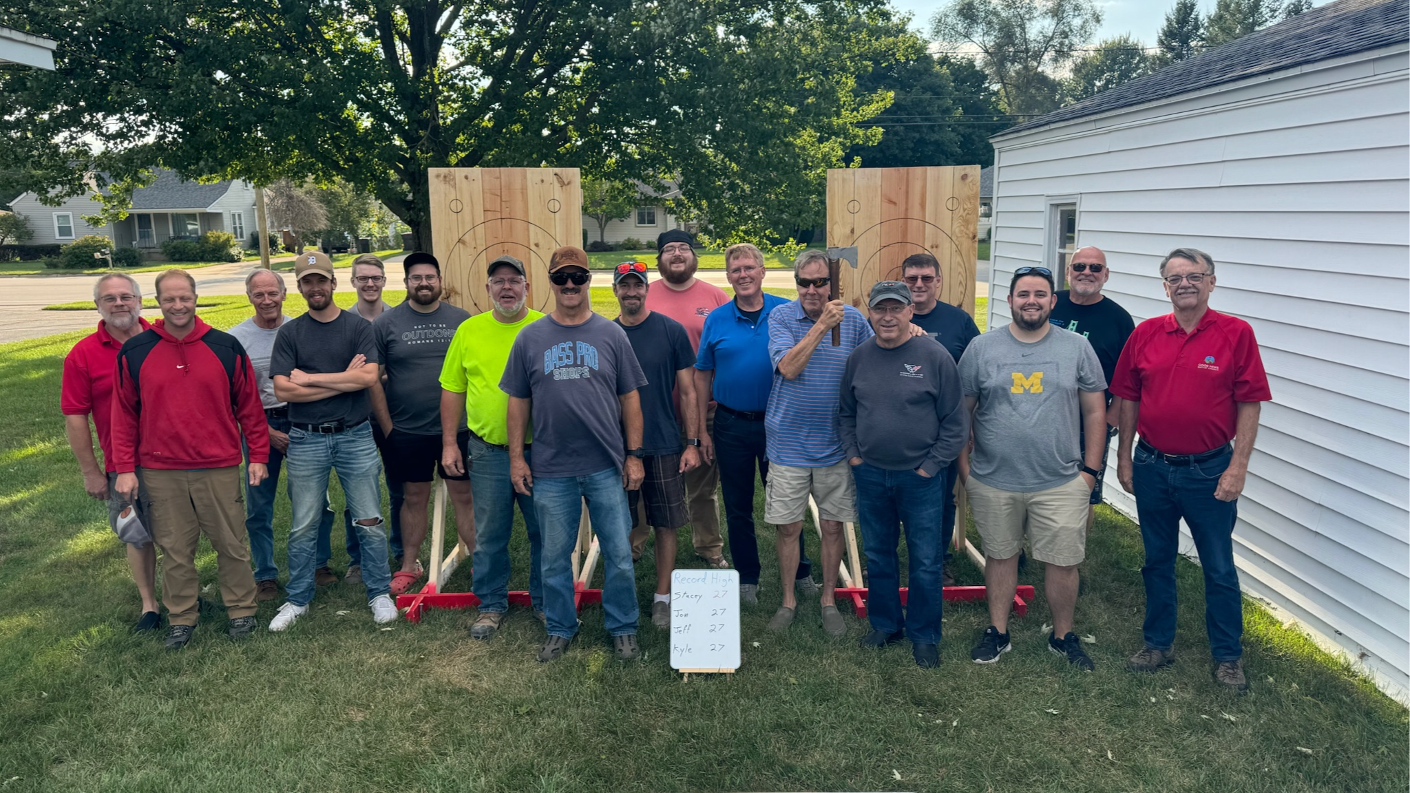 Good News Baptist Church Grand Rapids Michigan Group of 16 men standing outdoors in front of wooden axe-throwing boards, with a white board showing scores and a sign in the grass. Trees and houses are in the background.