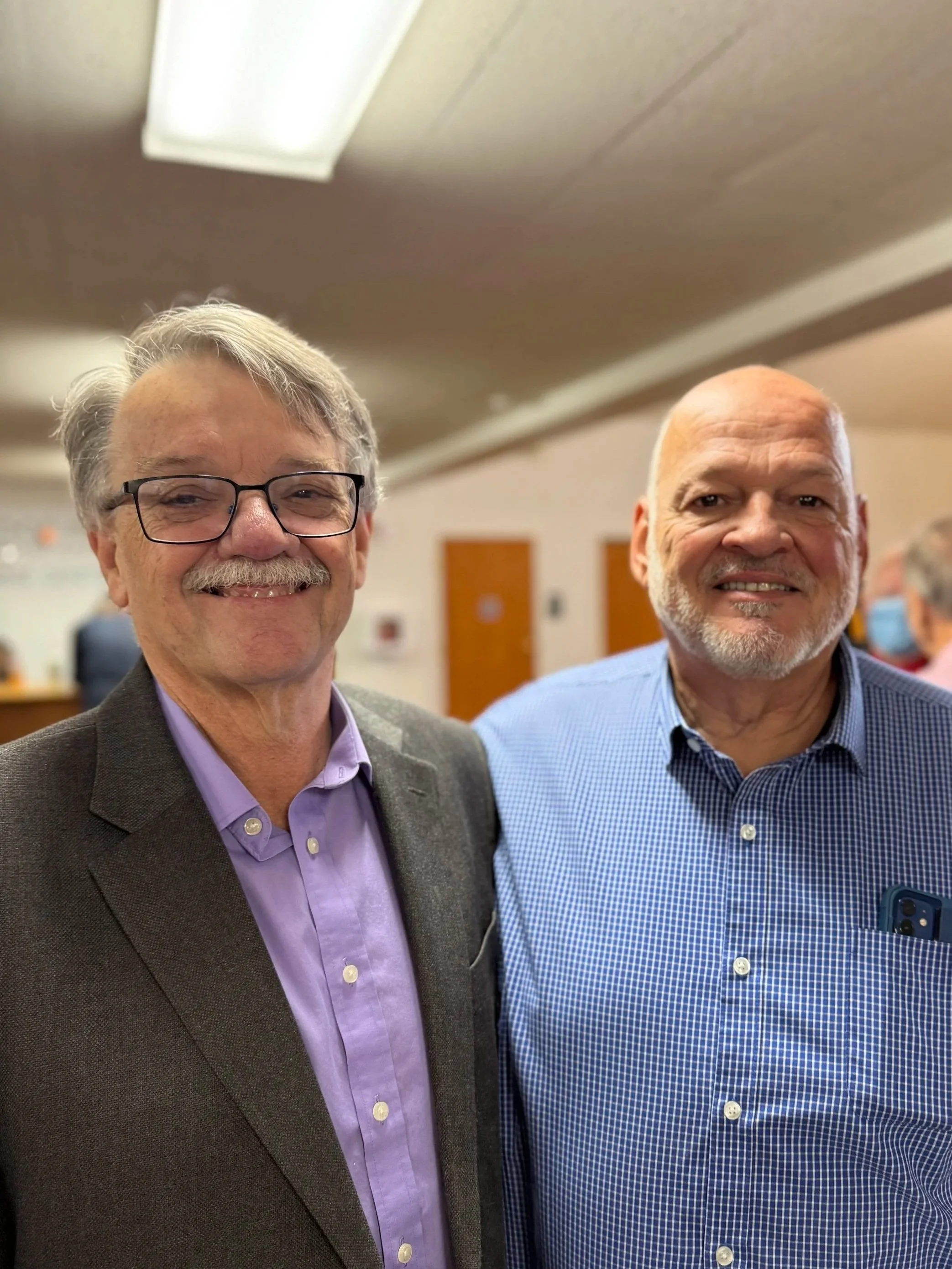 Good News Baptist Church Grand Rapids smiling men pose together indoors; one has glasses and a mustache, wearing a dark blazer and light purple shirt, the other is bald with a gray beard, wearing a blue checkered shirt with a smartphone in pocket