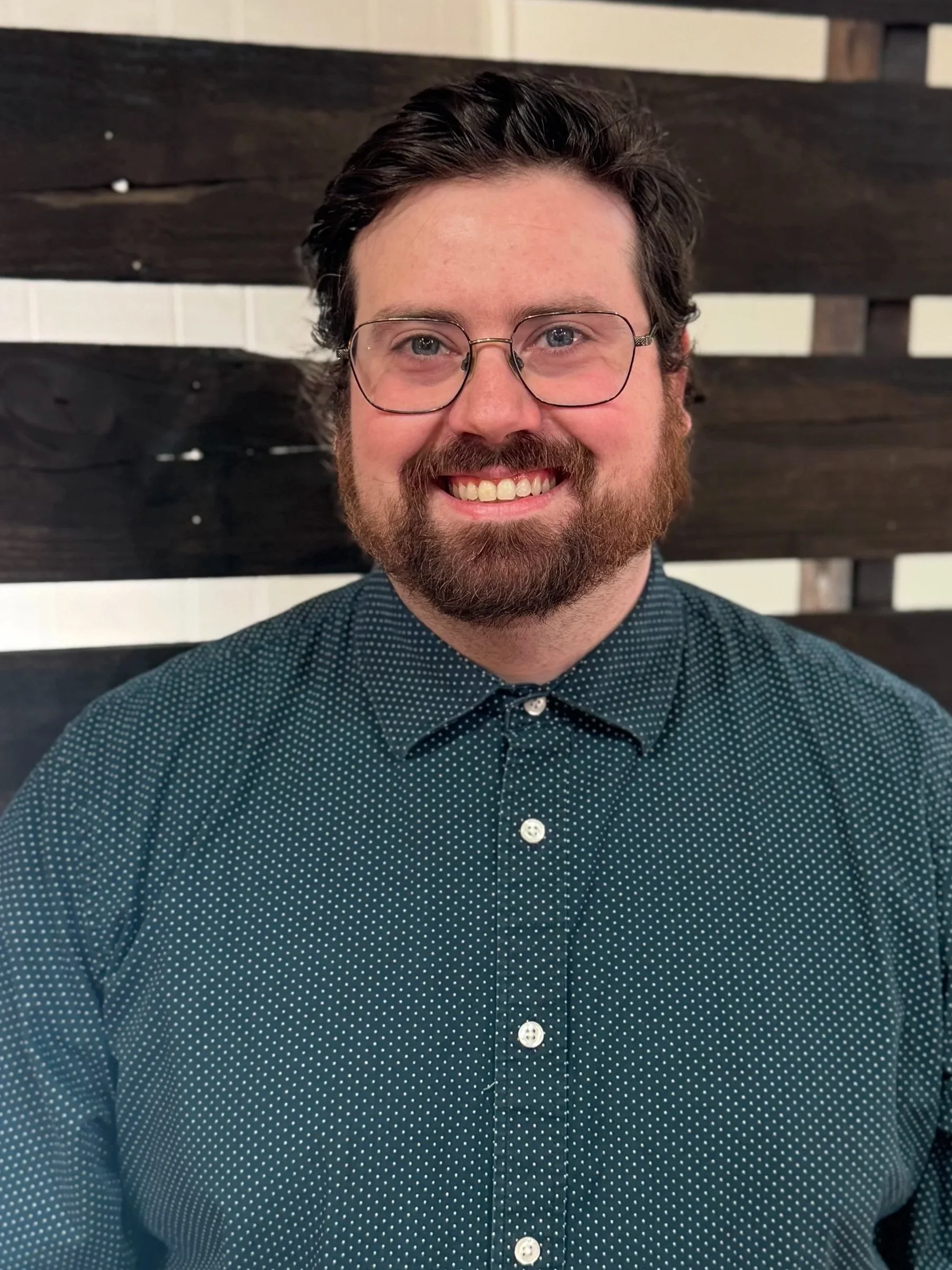 Good News Baptist Church Grand Rapids Michigan A man with glasses, a beard, and dark hair smiling at the camera. He is wearing a dark green, polka-dotted shirt and standing in front of a decorative wooden wall.