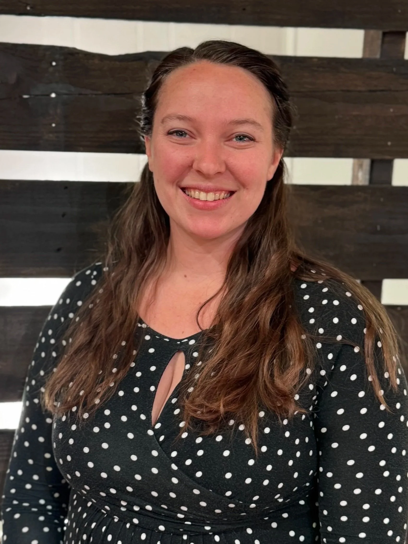 Good News Baptist Church Grand Rapids Michigan A woman with long wavy brown hair, wearing a black dress with white polka dots and a keyhole neckline, is smiling and standing in front of a wooden slat wall.