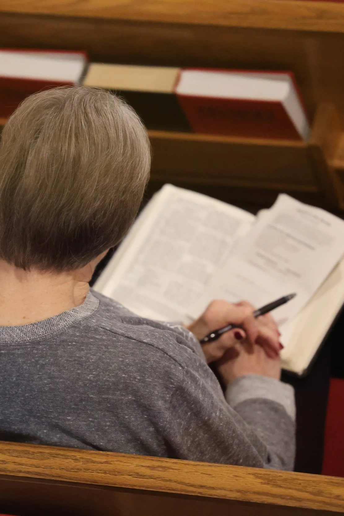 Good News Baptist Church Grand Rapids Michigan A person with short gray hair reading a book or Bible with a pen in hand, seated at a wooden pew with red and white books on a shelf behind them.