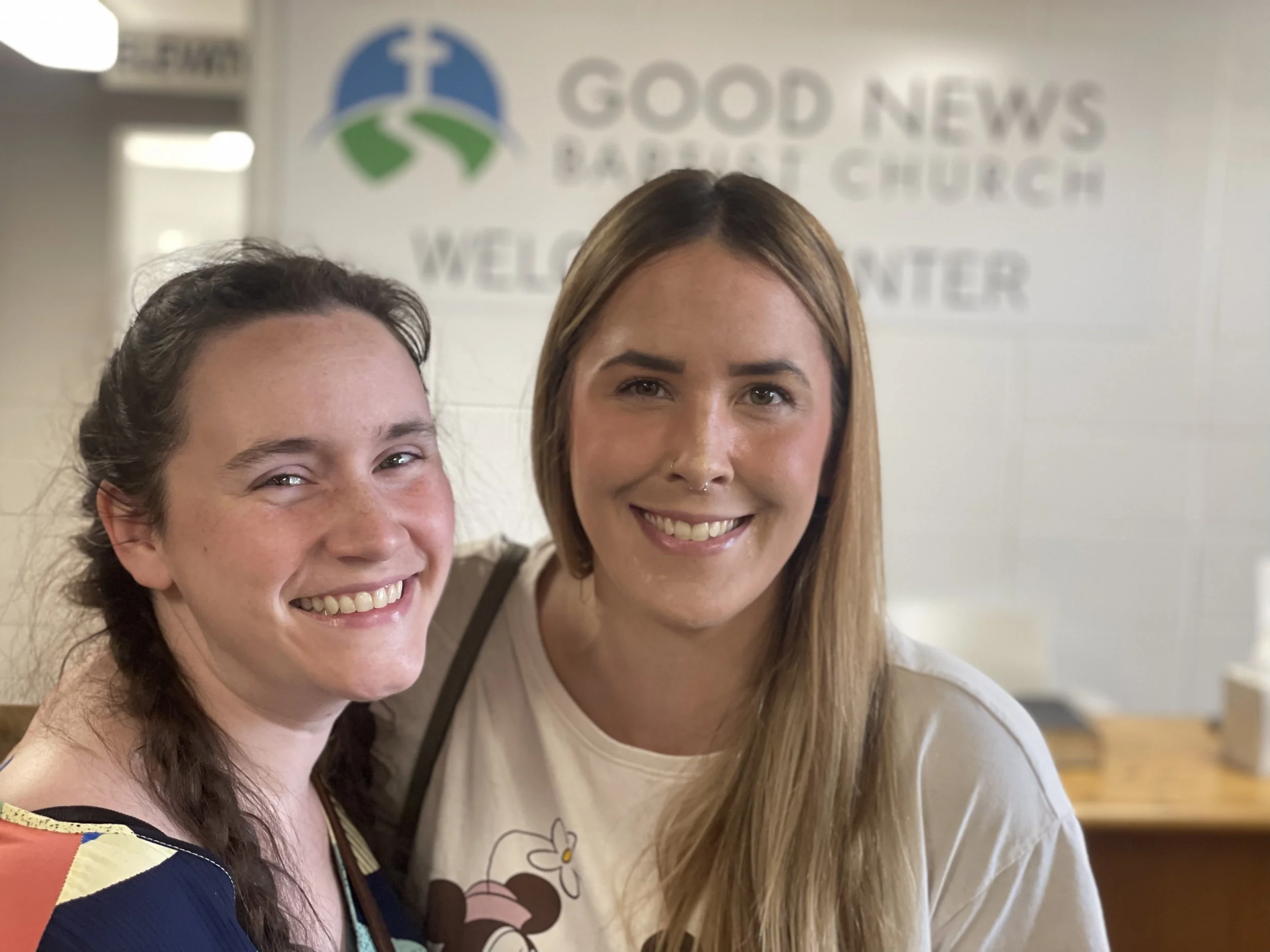Two women smiling in front of a sign for Good News Baptist Church Grand Rapids Mi. Cutlerville Gaines Township