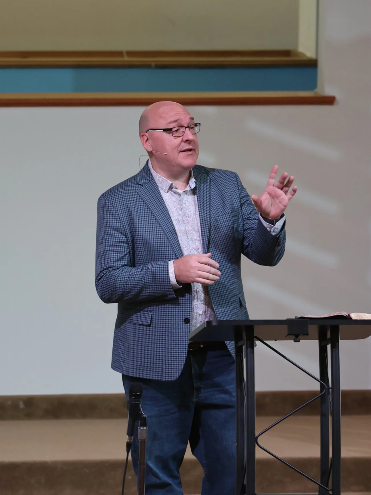 Good News Baptist Church Grand Rapids Michigan Pastor Brian Cederquist, wearing a checkered blazer, stands at a podium, speaking into a microphone, with a beige wall and blue architectural detail in the background.