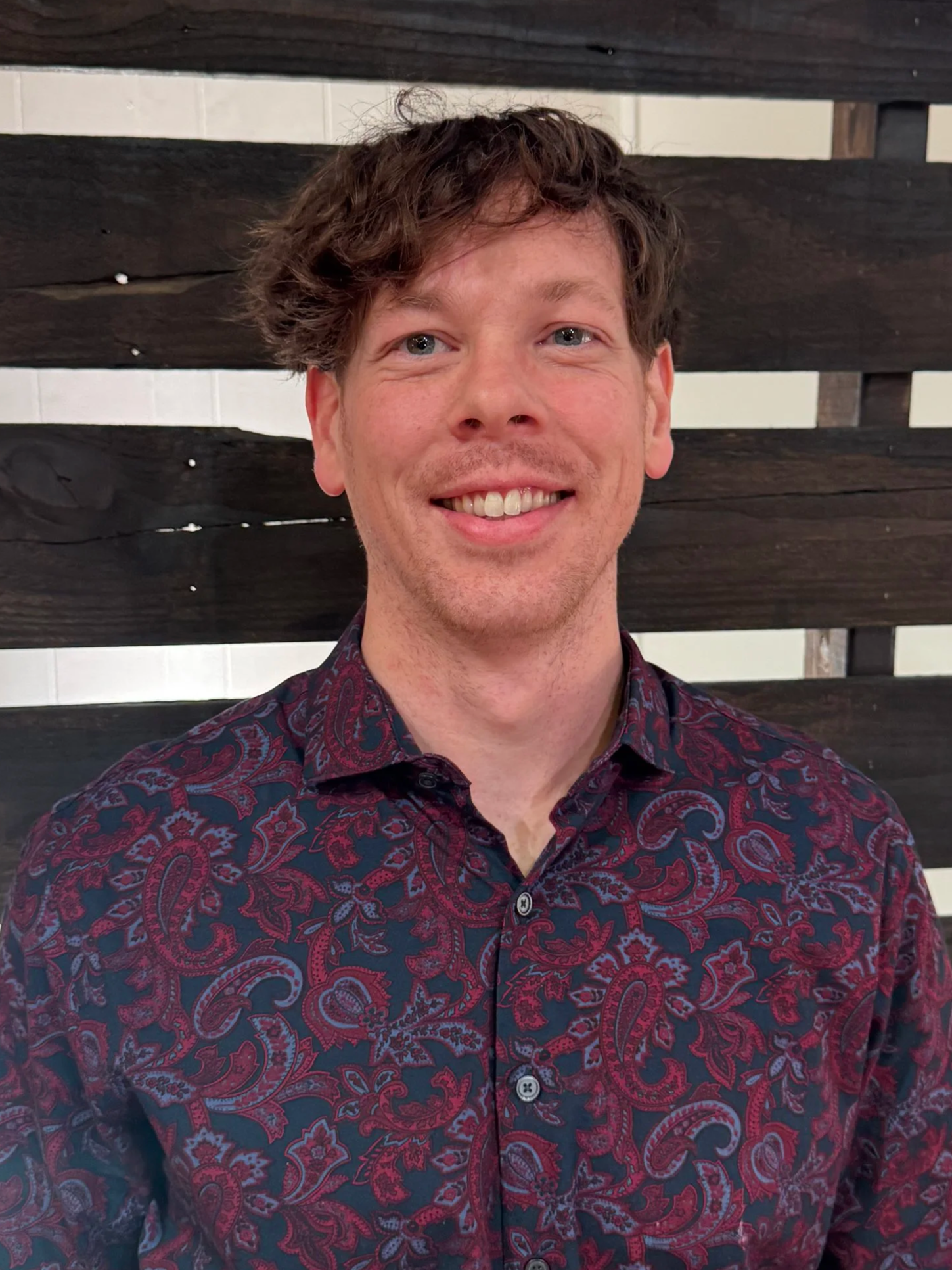 Good News Baptist Church Grand Rapids Michigan A smiling young man with brown hair wearing a dark patterned shirt, standing indoors in front of a wooden panel wall.