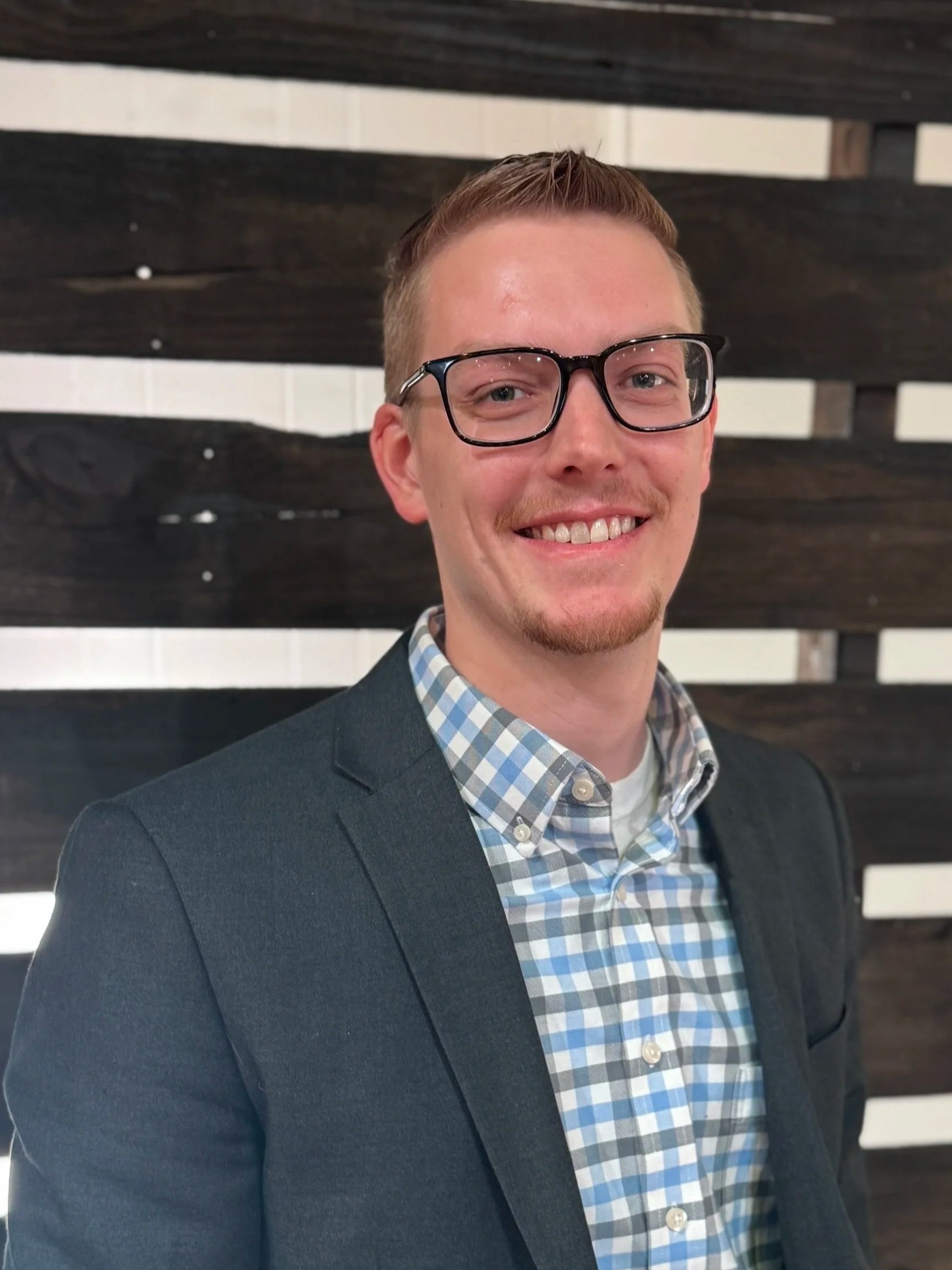 Good News Baptist Church Grand Rapids Michigan A young man with short hair, glasses, and a goatee smiling in front of a dark wooden wall, wearing a checkered shirt and a dark blazer.