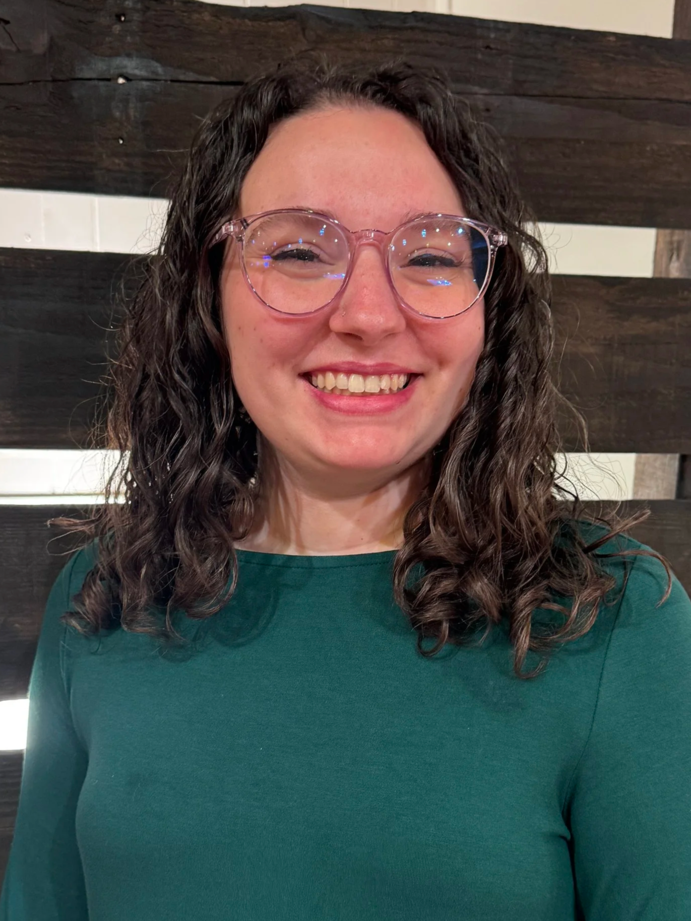 Good News Baptist Church Grand Rapids Michigan A woman with curly dark hair and glasses smiling in front of a dark wooden slat wall.