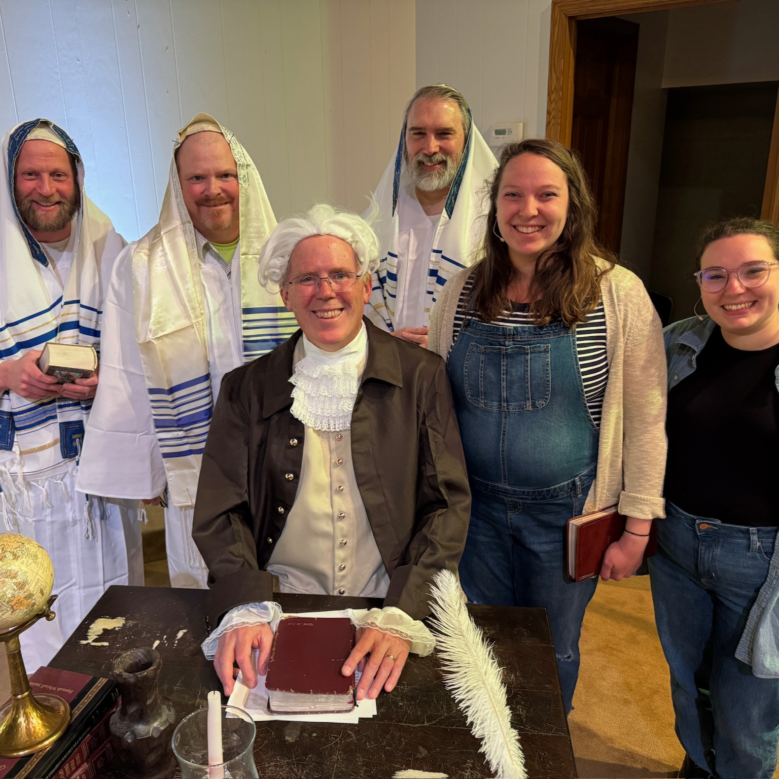 Good News Baptist Church Grand Rapids Michigan Group of people dressed in biblical costumes, with one man wearing traditional Jewish attire, gathered around a desk with a large book, a candle, and a white feather