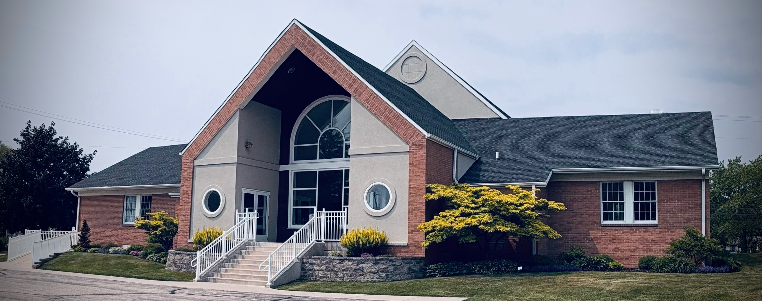 Good News Baptist Church Grand Rapids Michigan A modern church with brick and stucco exterior, large arched window, small circular windows, and a landscaped front yard with shrubs and trees.