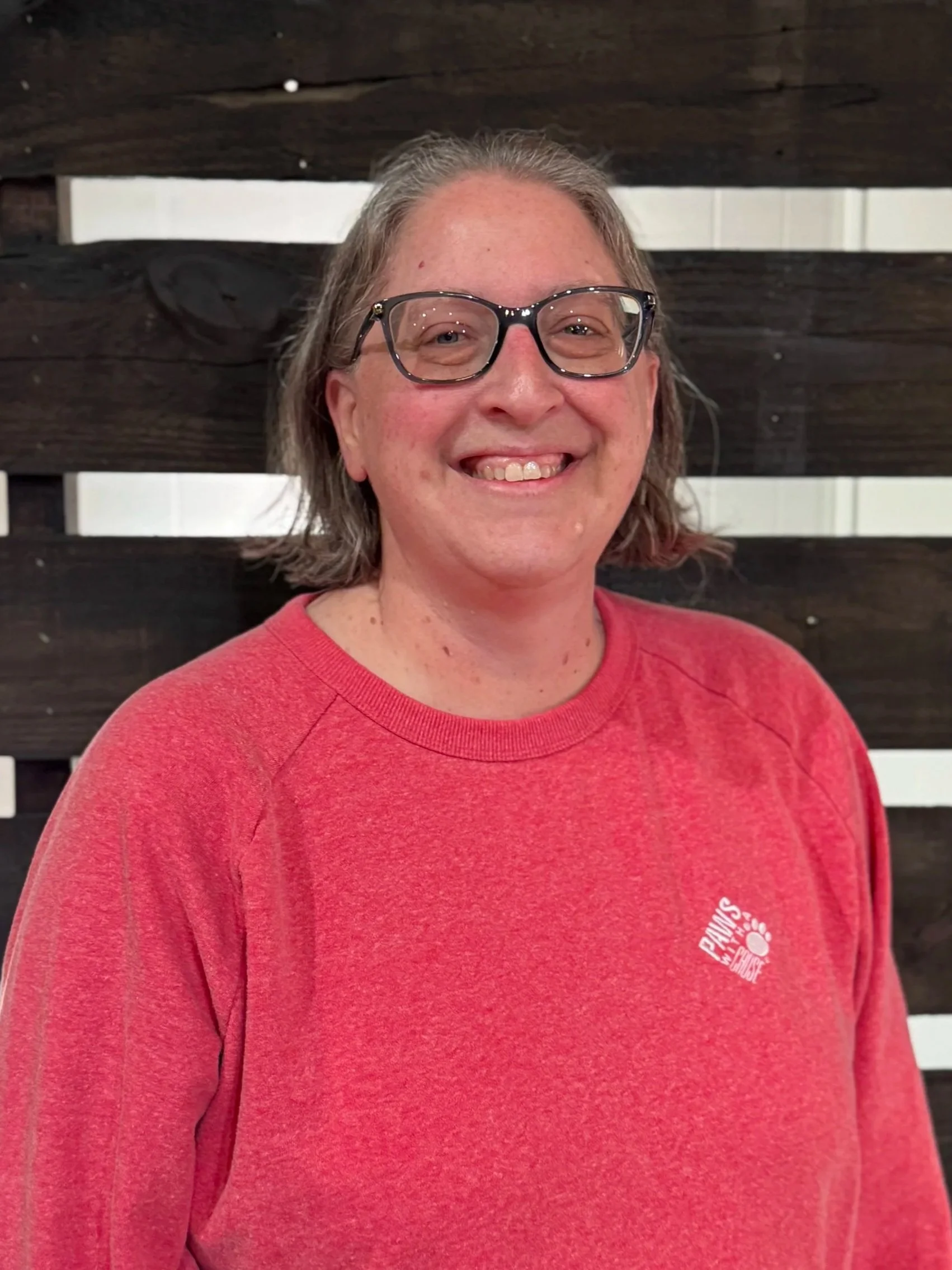 Good News Baptist Church Grand Rapids Michigan A woman with shoulder-length gray hair and glasses smiling, wearing a red sweatshirt, standing in front of a dark wooden fence.
