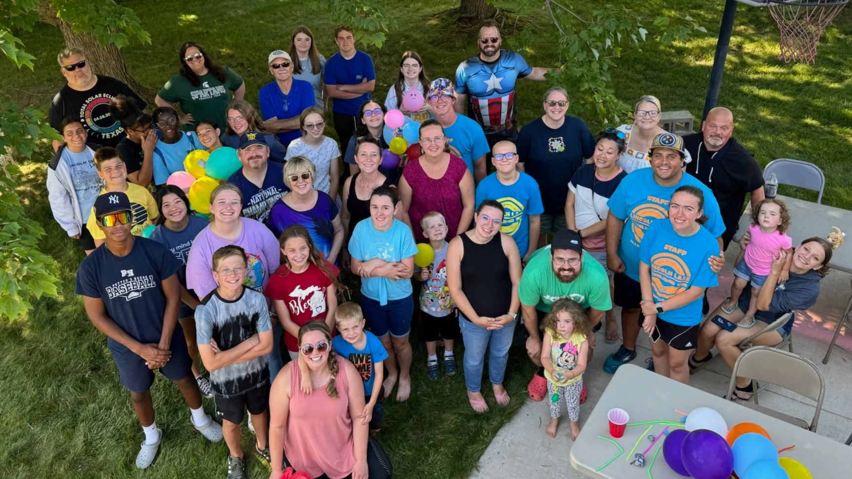 Good News Baptist Church Grand Rapids Michigan Community gathering of diverse children and adults outdoors with balloons, some wearing sports jerseys and casual clothes, under trees on a sunny day.