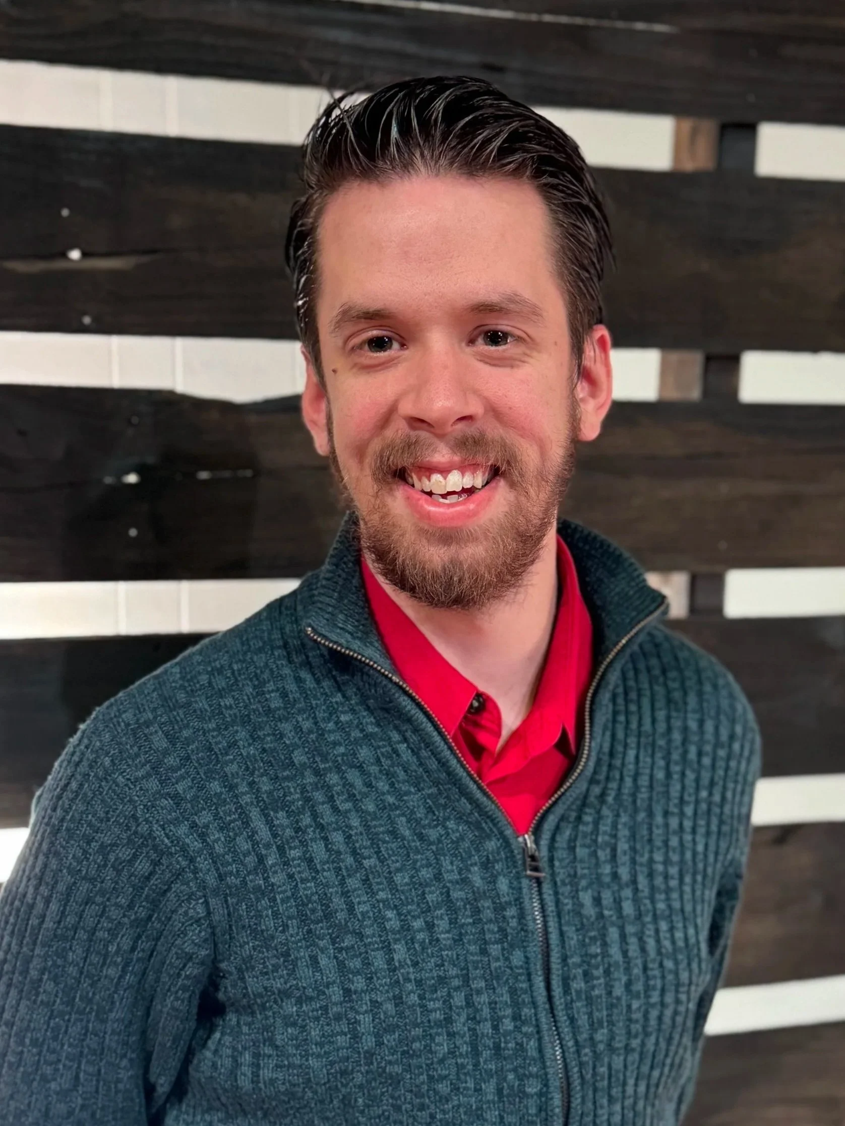 Good News Baptist Church Grand Rapids Michigan A smiling man with a beard and mustache, wearing a red shirt and a dark green zip-up sweater, standing in front of a modern wooden slat wall.