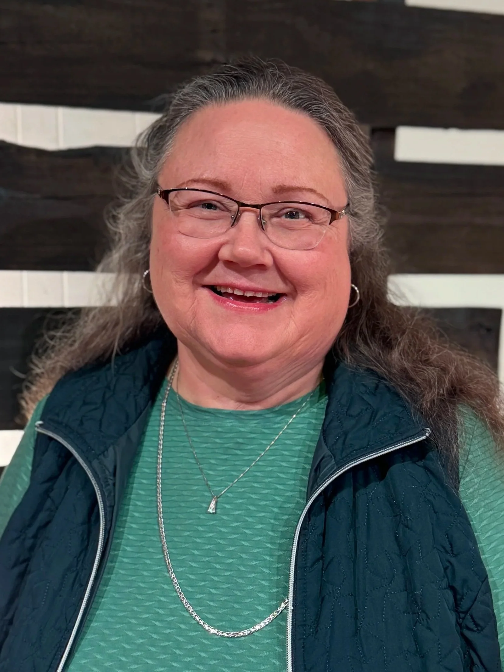 Good News Baptist Church Grand Rapids Michigan A smiling woman with glasses, long gray hair, wearing a green textured shirt, a navy quilted vest, and layered silver necklaces, standing in front of a decorative wooden wall.