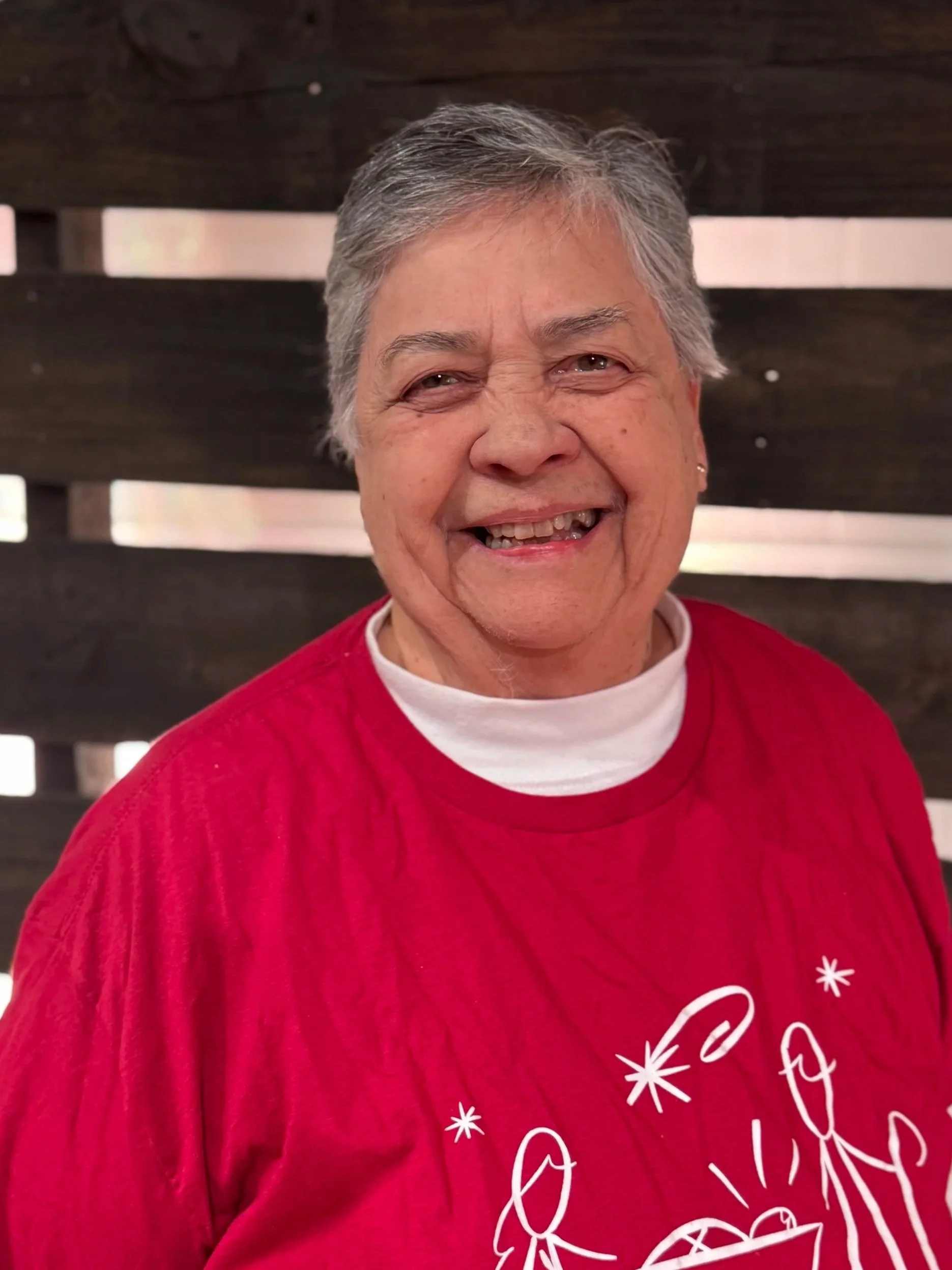 Good News Baptist Church Grand Rapids Michigan Smiling elderly woman wearing a red Christmas T-shirt with a nativity scene and the text 'Teach the Children' standing in front of a dark wooden fence.