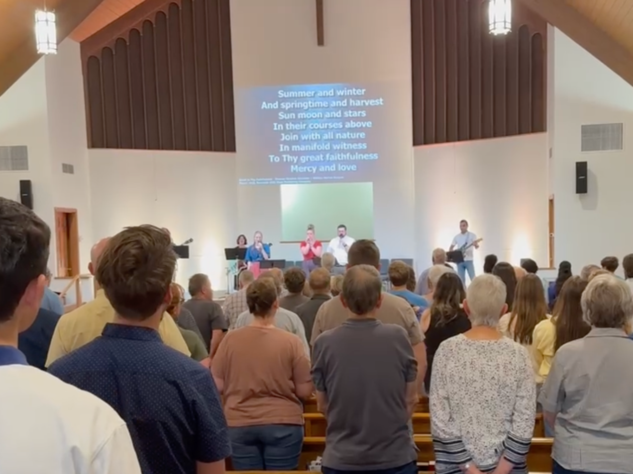 Good News Baptist Church Grand Rapids Michigan - A congregation gathered in a church during a worship service, with five people leading worship at the front, including singers and musicians, while the audience stands facing the stage.