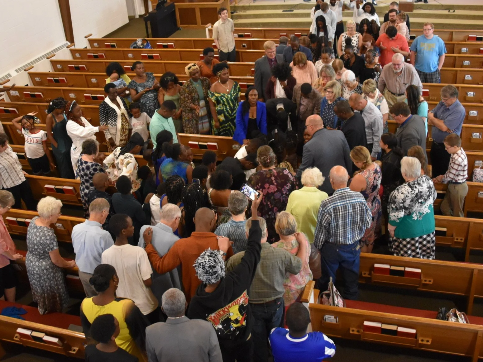 Good News Baptist Church Grand Rapids Michigan A diverse group of people gathered inside a church, some standing in aisles and others seated on wooden pews, participating in a prayer or moment of reflection.