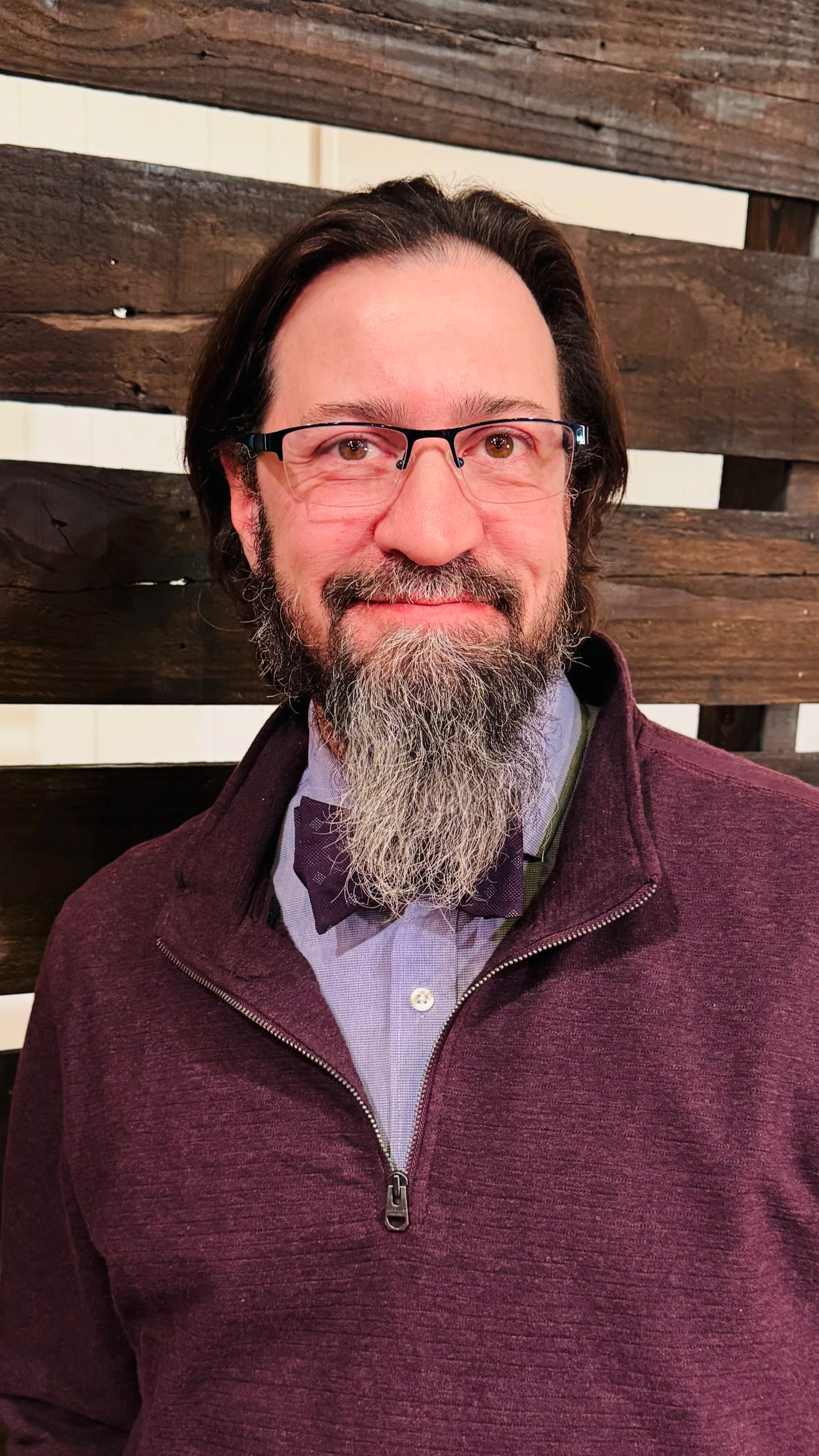 Good News Baptist Church Grand Rapids Michigan Man with glasses, a beard, and a bow tie, wearing a maroon quarter-zip sweater, standing in front of a wooden wall.