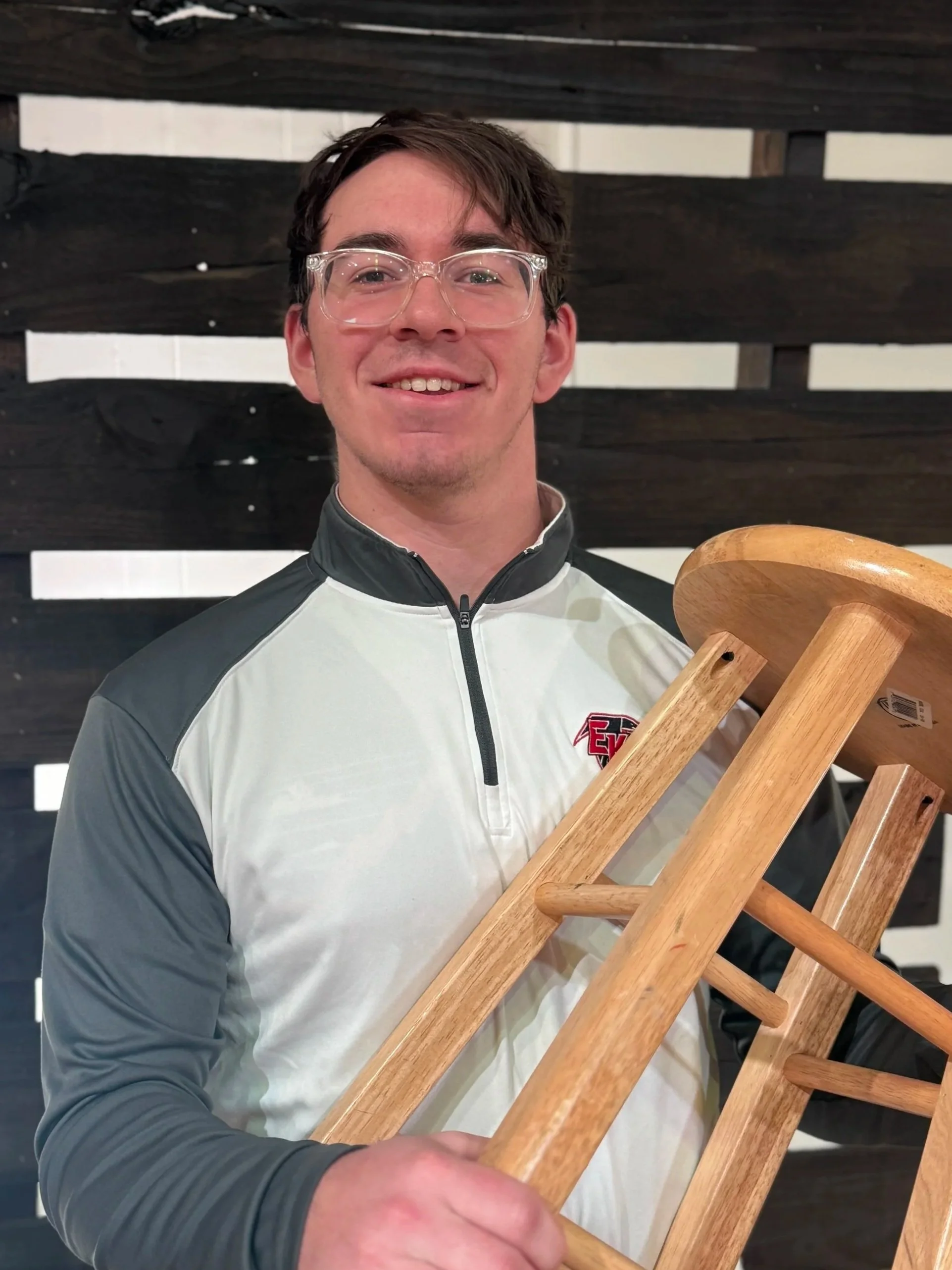 Good News Baptist Church Grand Rapids Michigan A young man smiling, wearing glasses and a jacket, holding a wooden stool in a room with a black wooden wall in the background.