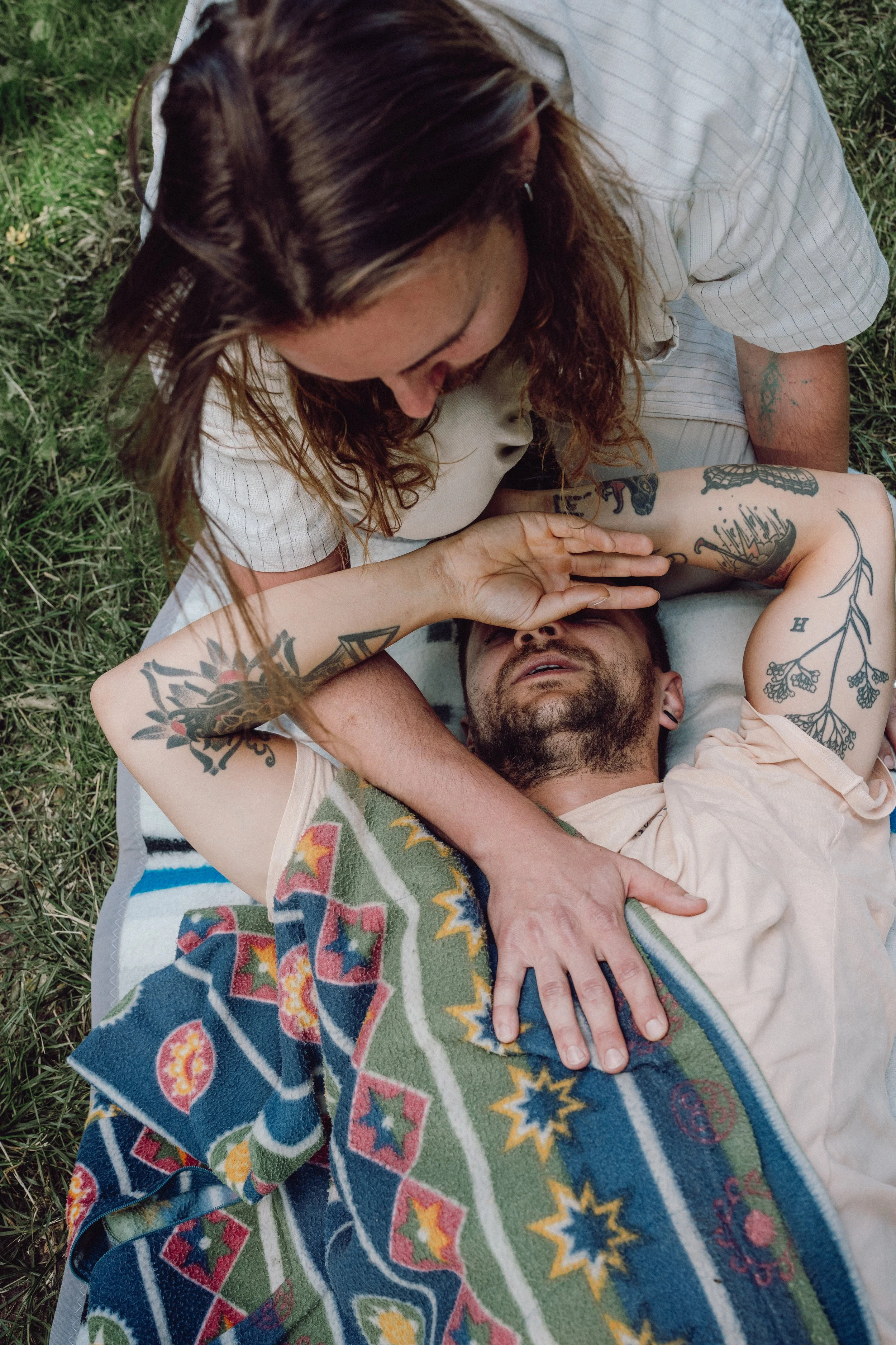 Ein Mann liegt auf einer Decke im Gras, während eine Frau über ihm steht und ihm ins Gesicht schaut. Sie berühren sich eine Hand und scheinen ein intimes Gespräch zu führen.