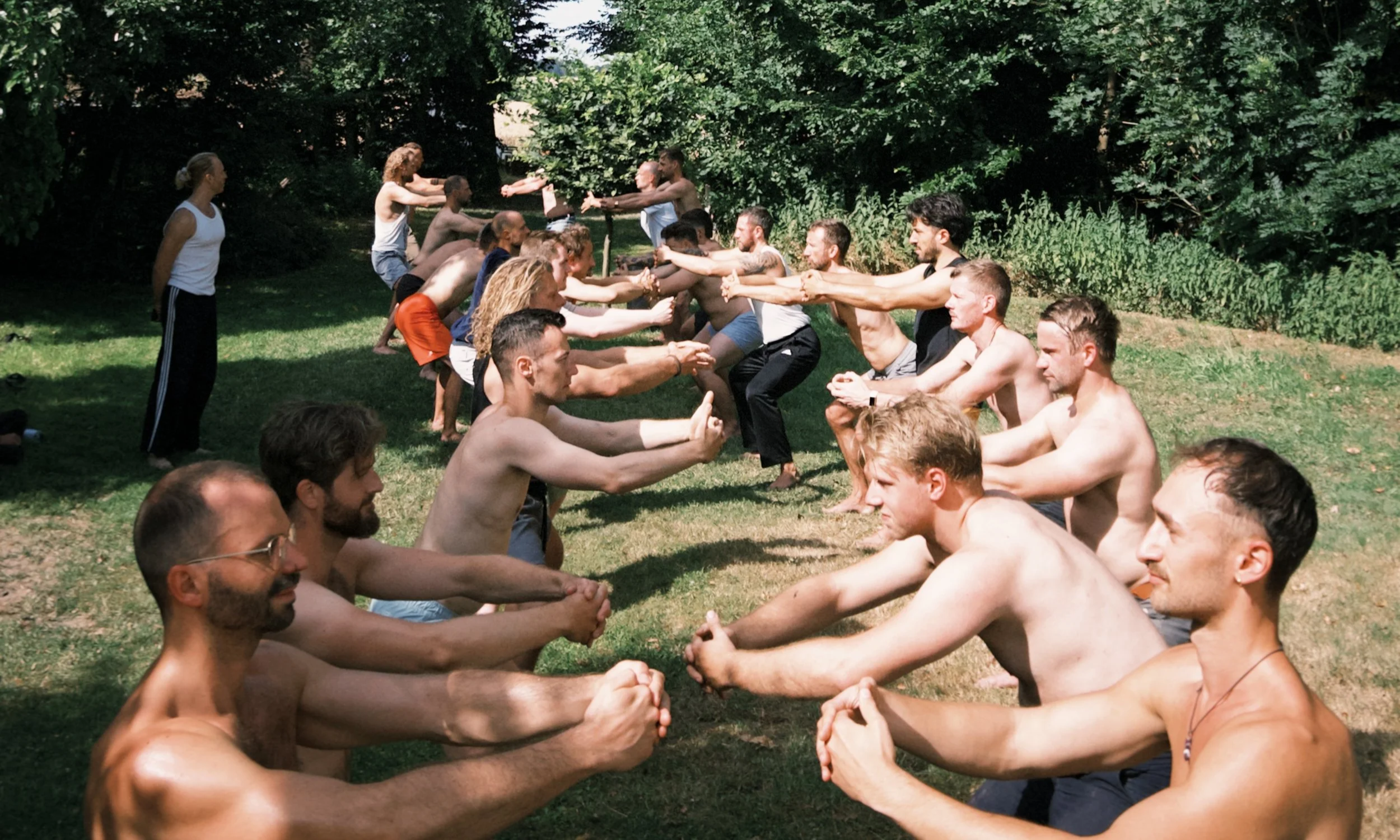 Gruppe von Menschen, die an einem Tafel im Freien Yoga oder Meditation üben, sitzend auf dem Gras, umgeben von Bäumen an einem sonnigen Tag.