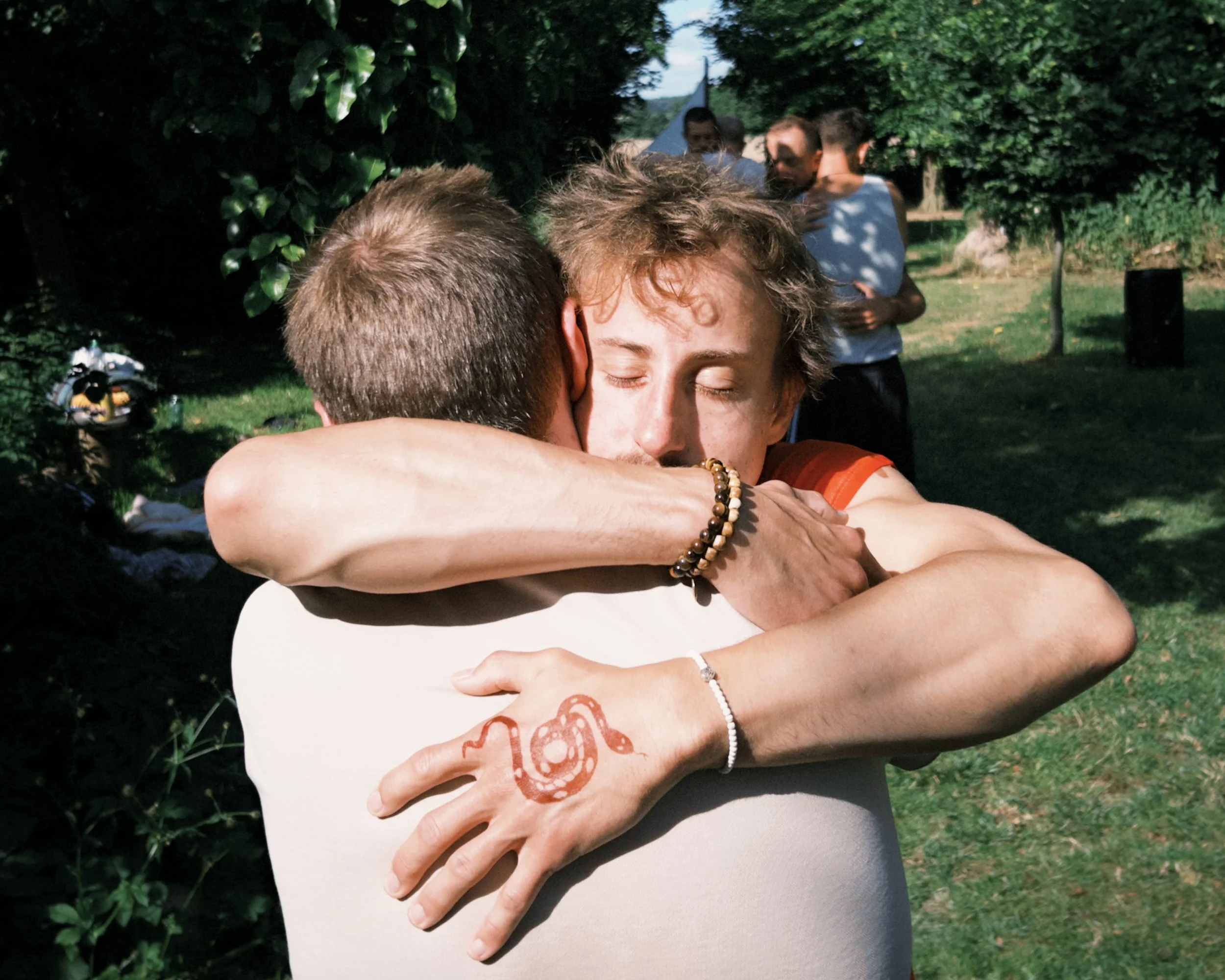Zwei Menschen umarmen sich im Freien, eine Person hat eine Henna-Tattoo auf der Hand, während andere im Hintergrund stehen.