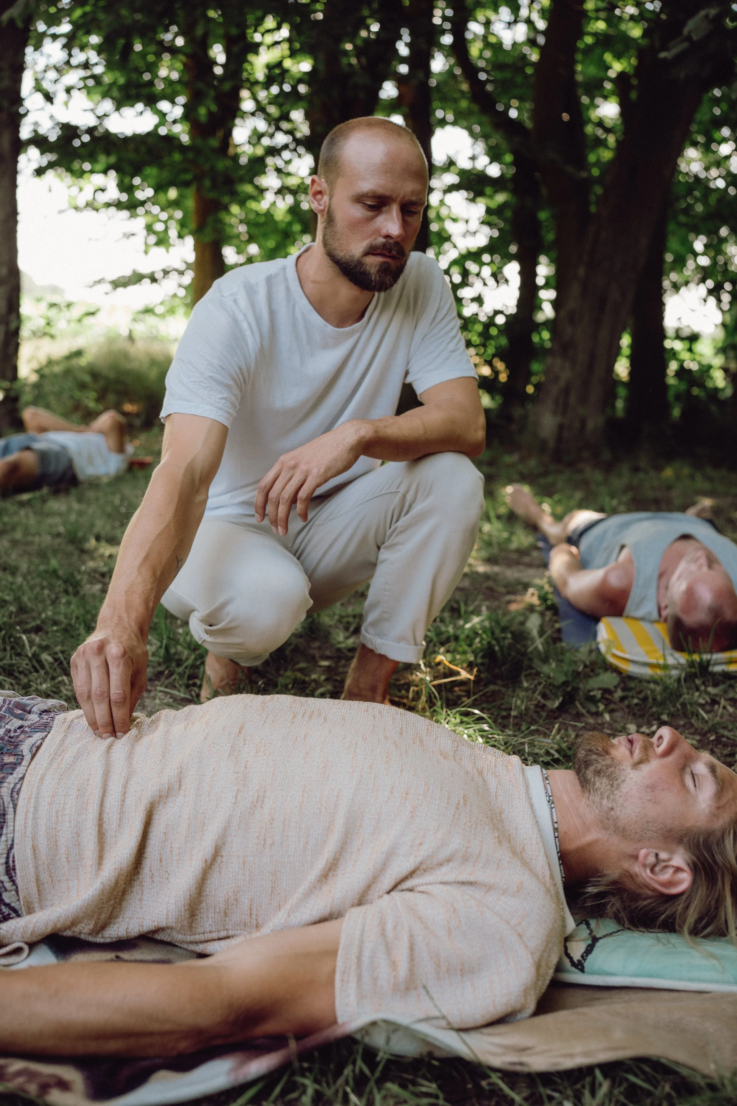 Ein Mann in weißen Kleidung, der in einem Wald kniet und auf eine Person, die bewusstlos auf der Erde liegt, schaut.
