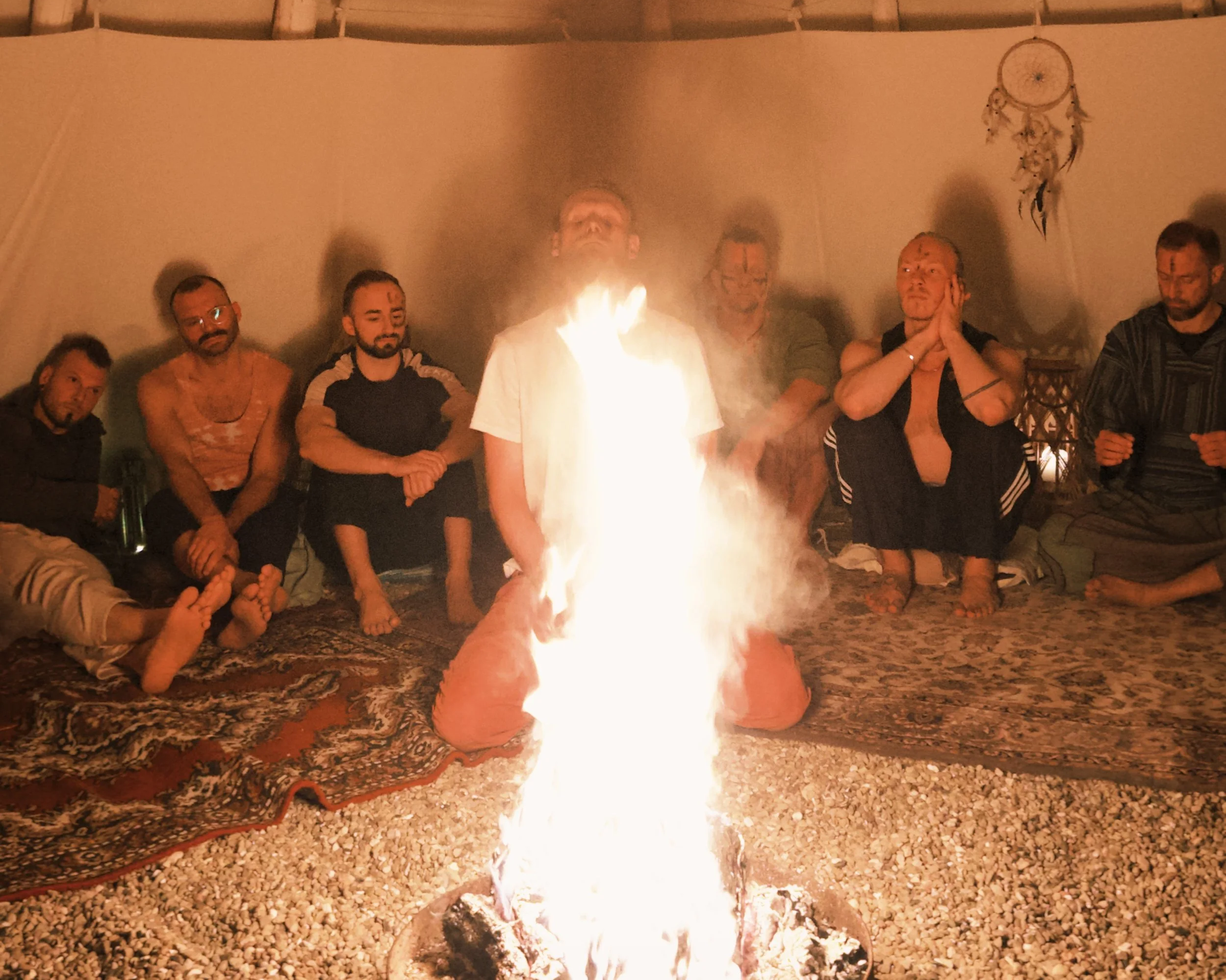 Menschen sitzen im Kreis um ein Feuer in einem Zelt, ein Mann scheint Feuer zu speien, alle sind entspannt und beobachten.