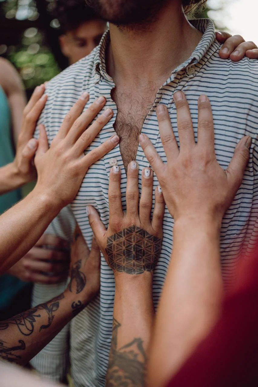 Mehrere Menschen berühren eine Person im grauen gestreiften Hemd an verschiedenen Stellen, was möglicherweise eine spirituelle oder rituelle Szene ist.