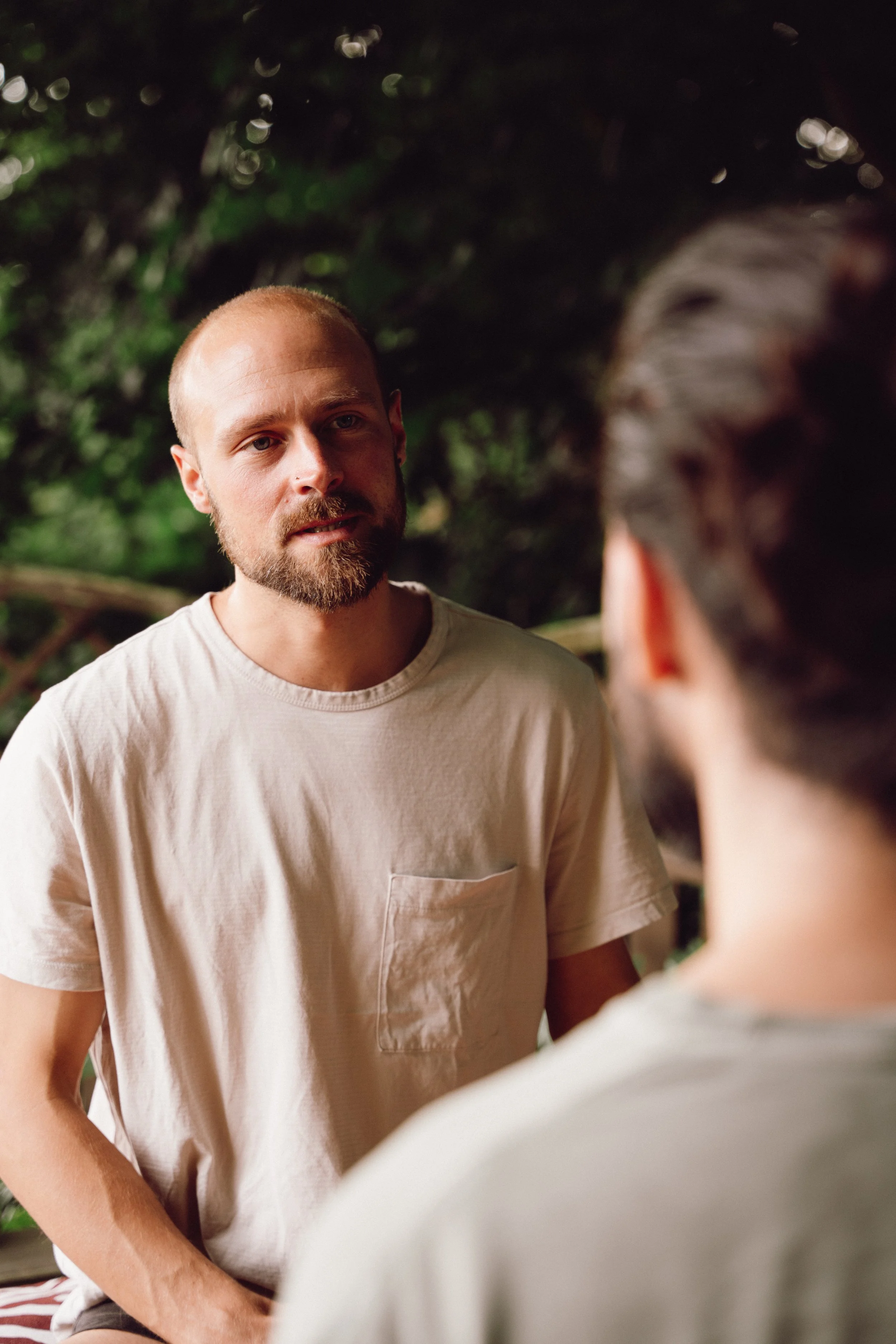 Zwei Männer im Gespräch in einem grünen, bewachsenen Umfeld, der Mann im Vordergrund trägt ein beige T-Shirt.