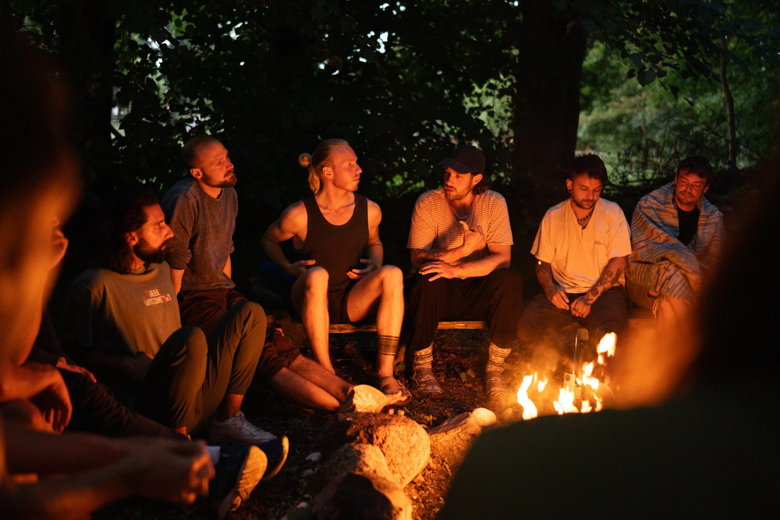 Gruppen von Menschen sitzen um ein Lagerfeuer im Wald, nachts, bei Gesprächen.