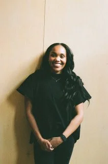 Portrait of a woman with long dark hair, smiling, wearing a black top against a neutral background.