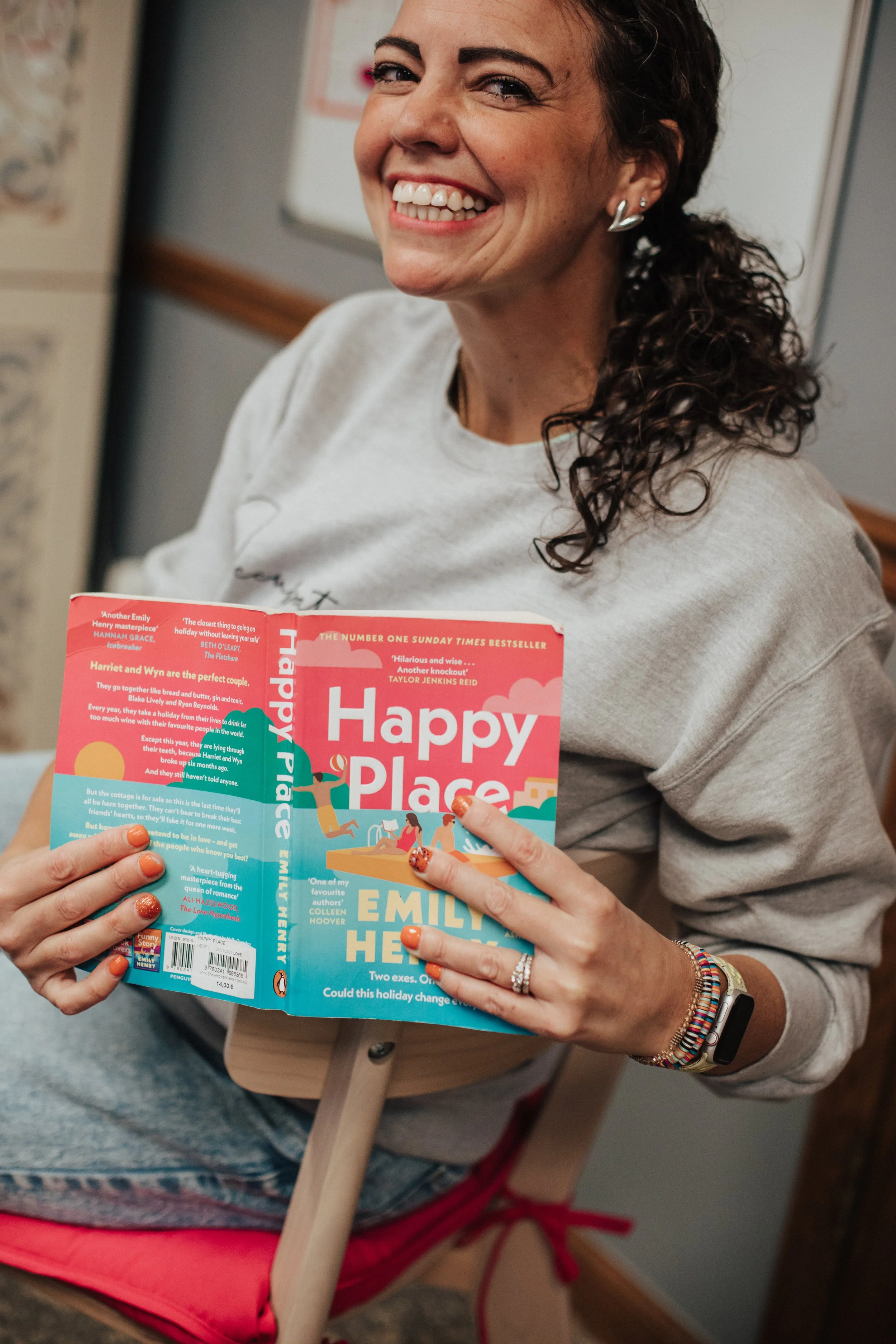 A woman with curly brown hair, smiling and holding a book titled 'Happy Place' by Emily Henry, inside a room with a whiteboard and artwork on the wall.
