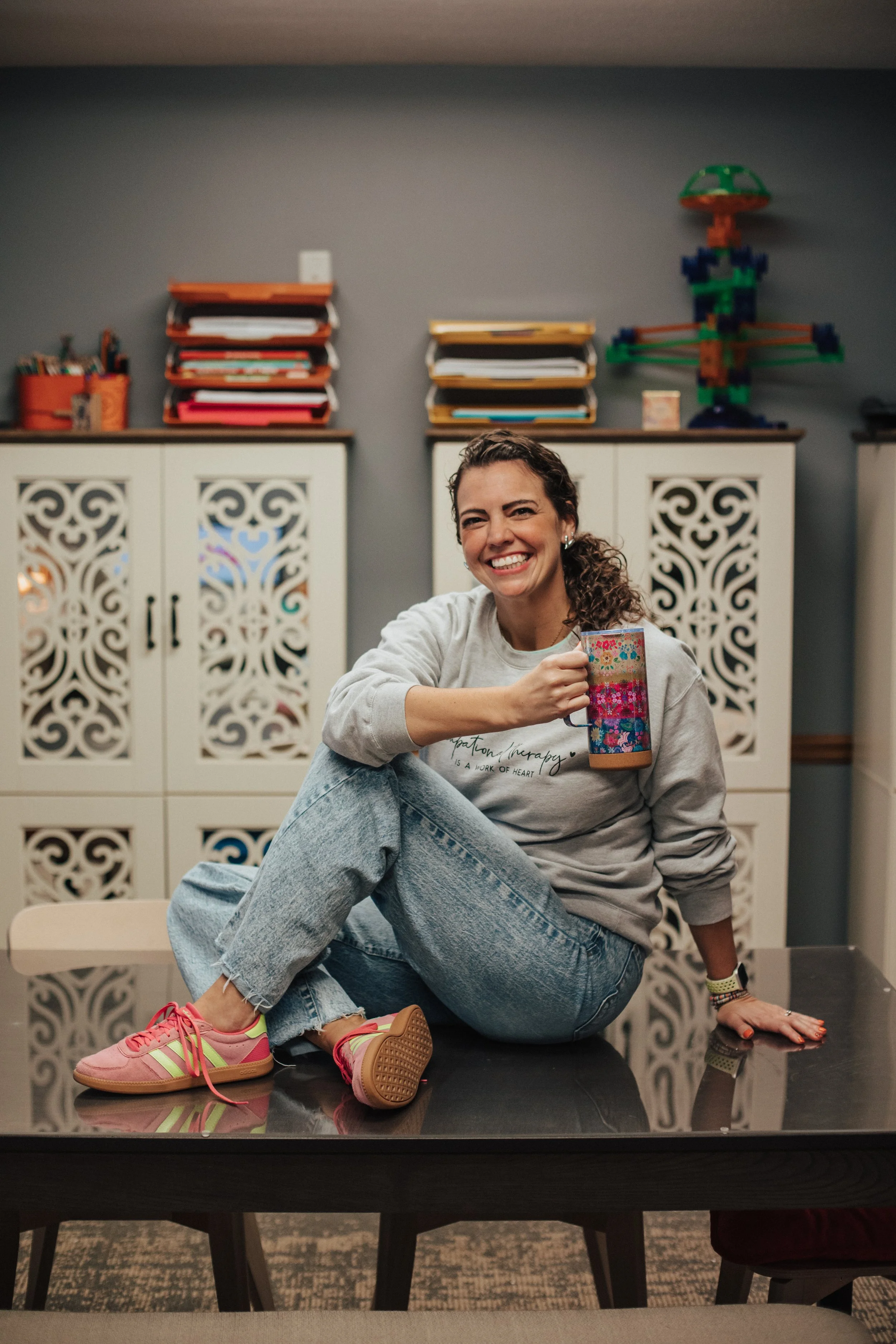 A woman sitting on a table, smiling and holding a colorful coffee mug in a room with storage cabinets and colorful paper stacks on top.