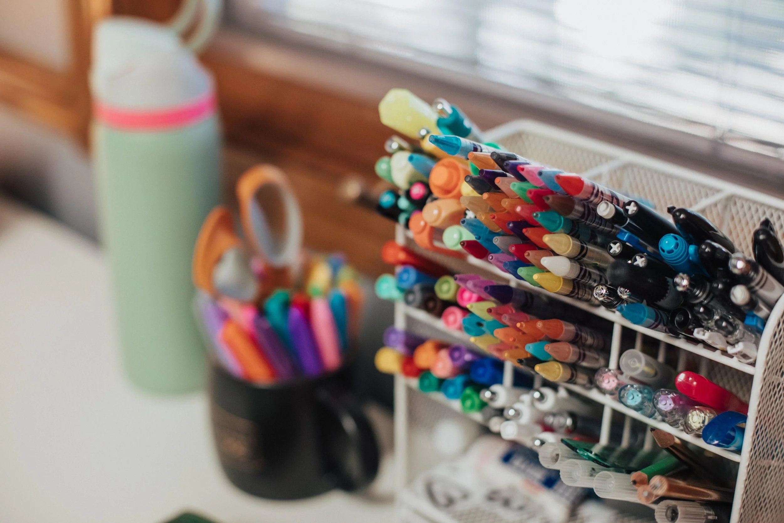Organized collection of colored markers, pens, and scissors on a desk near a window.