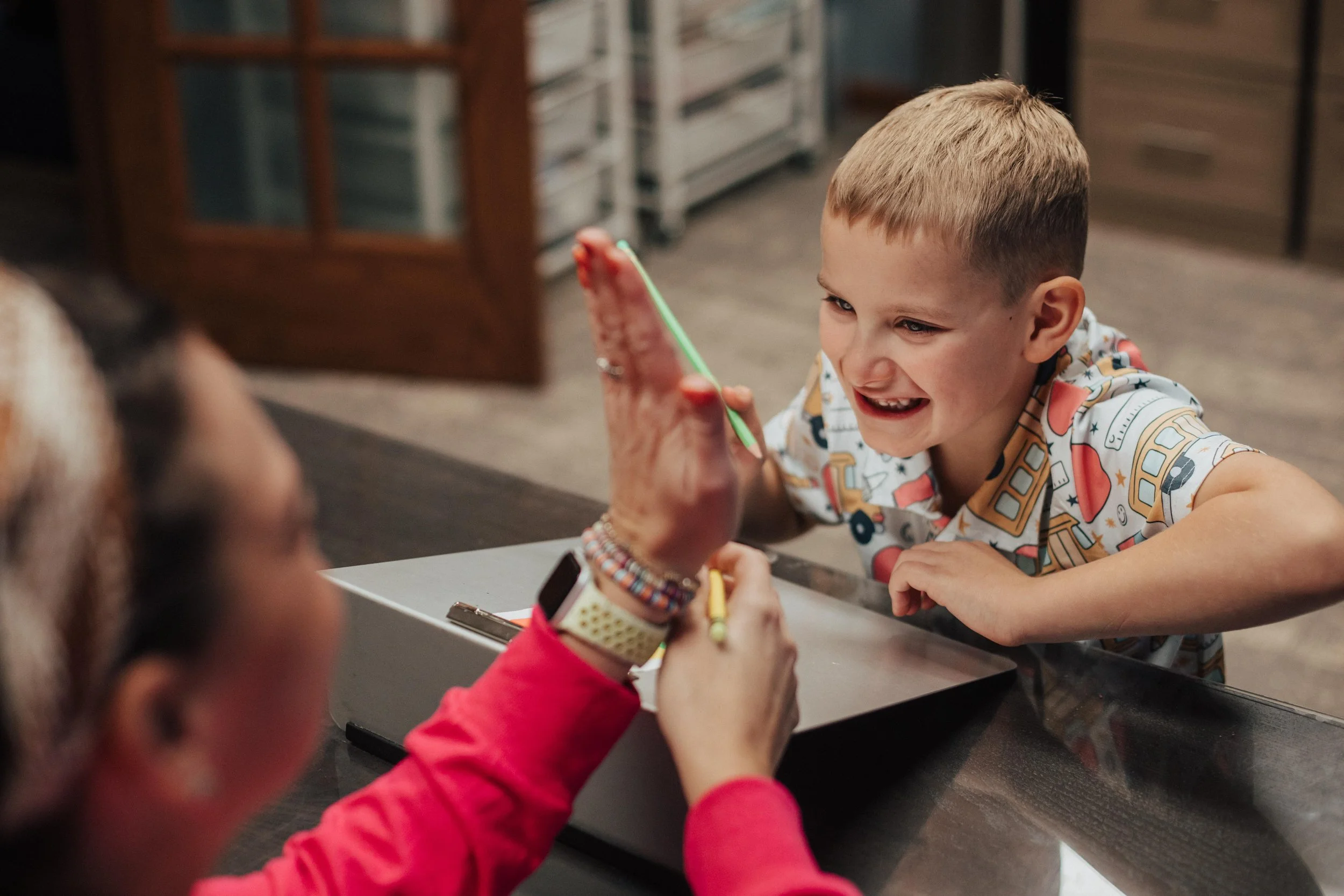 A young boy smiling and leaning over a table, reaching toward an occupational therapist in Westerville, OH who is giving him a high five.