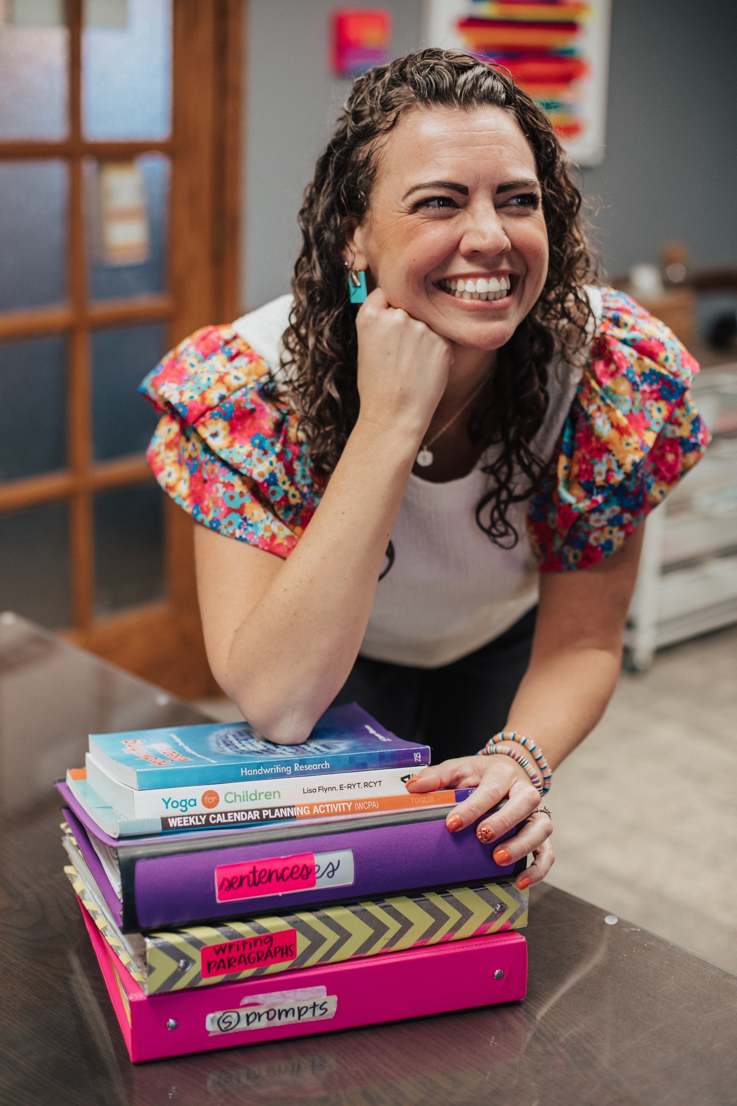 A woman smiling and leaning on a stack of colorful binders and notebooks on a table in a classroom setting