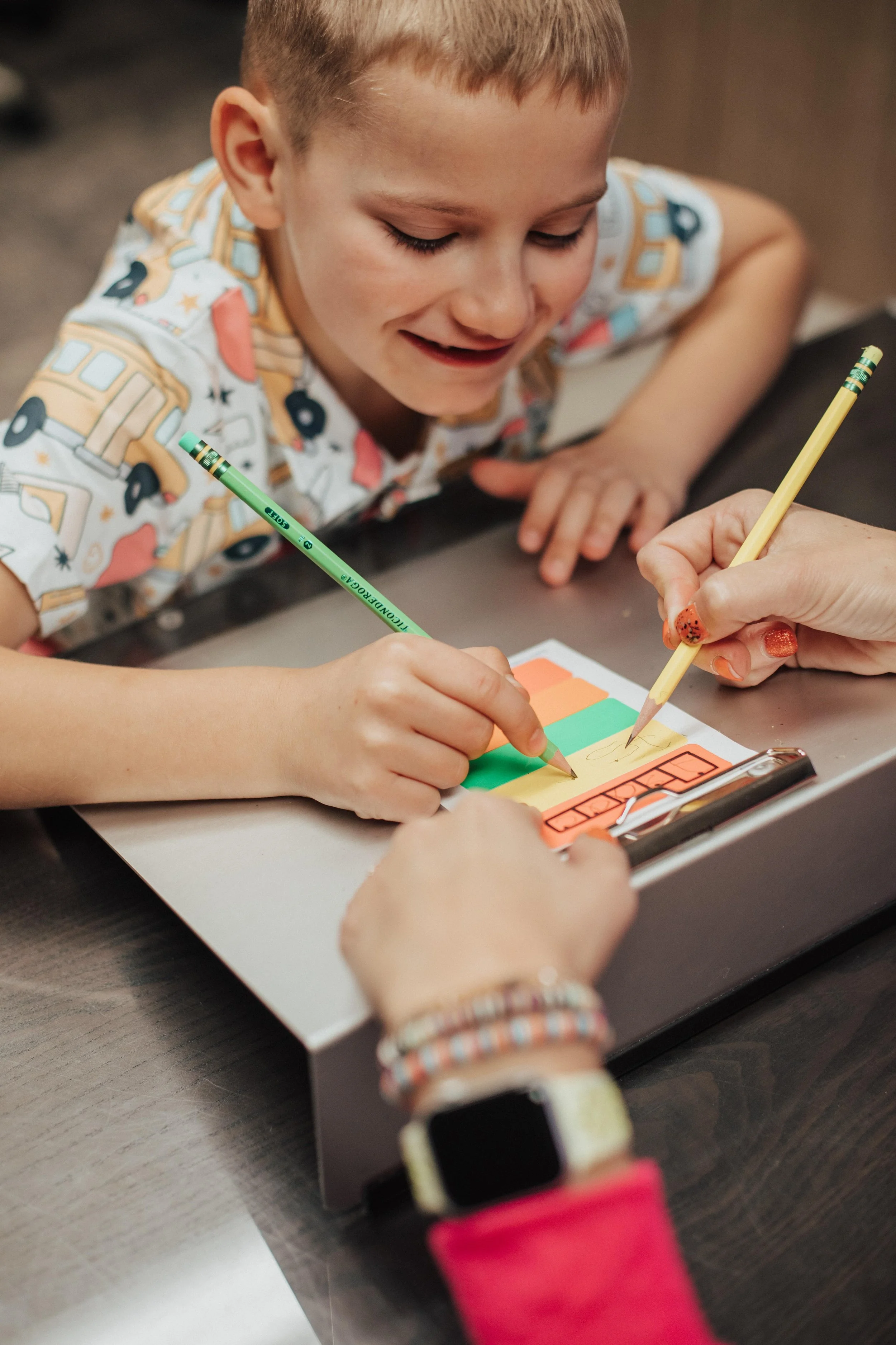 A young boy smiling as he writes on sticky notes at a table with the help of an adult, possibly in a classroom or office setting.