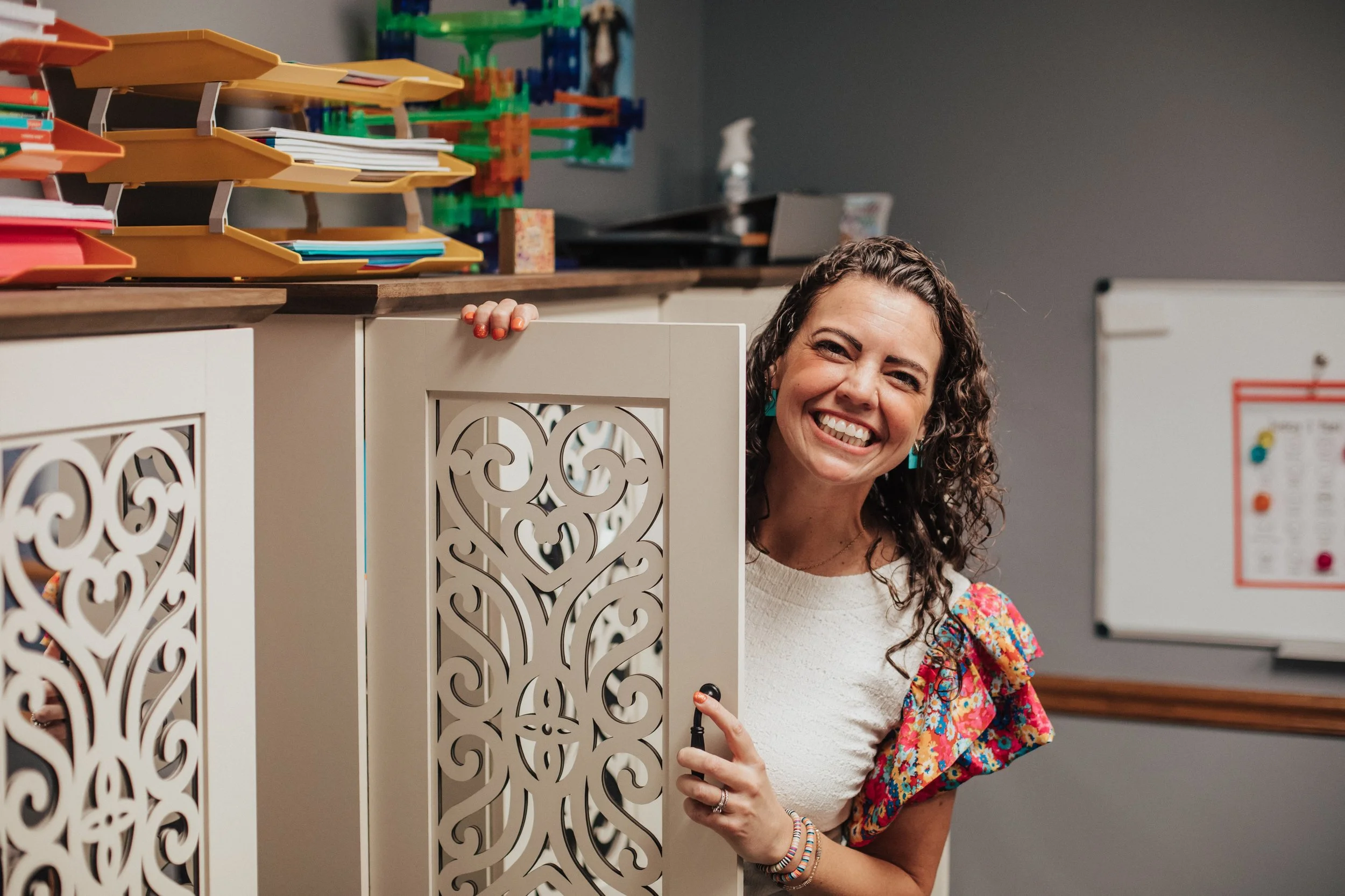 A woman smiling and peeking out from behind a decorative white cabinet in a classroom.